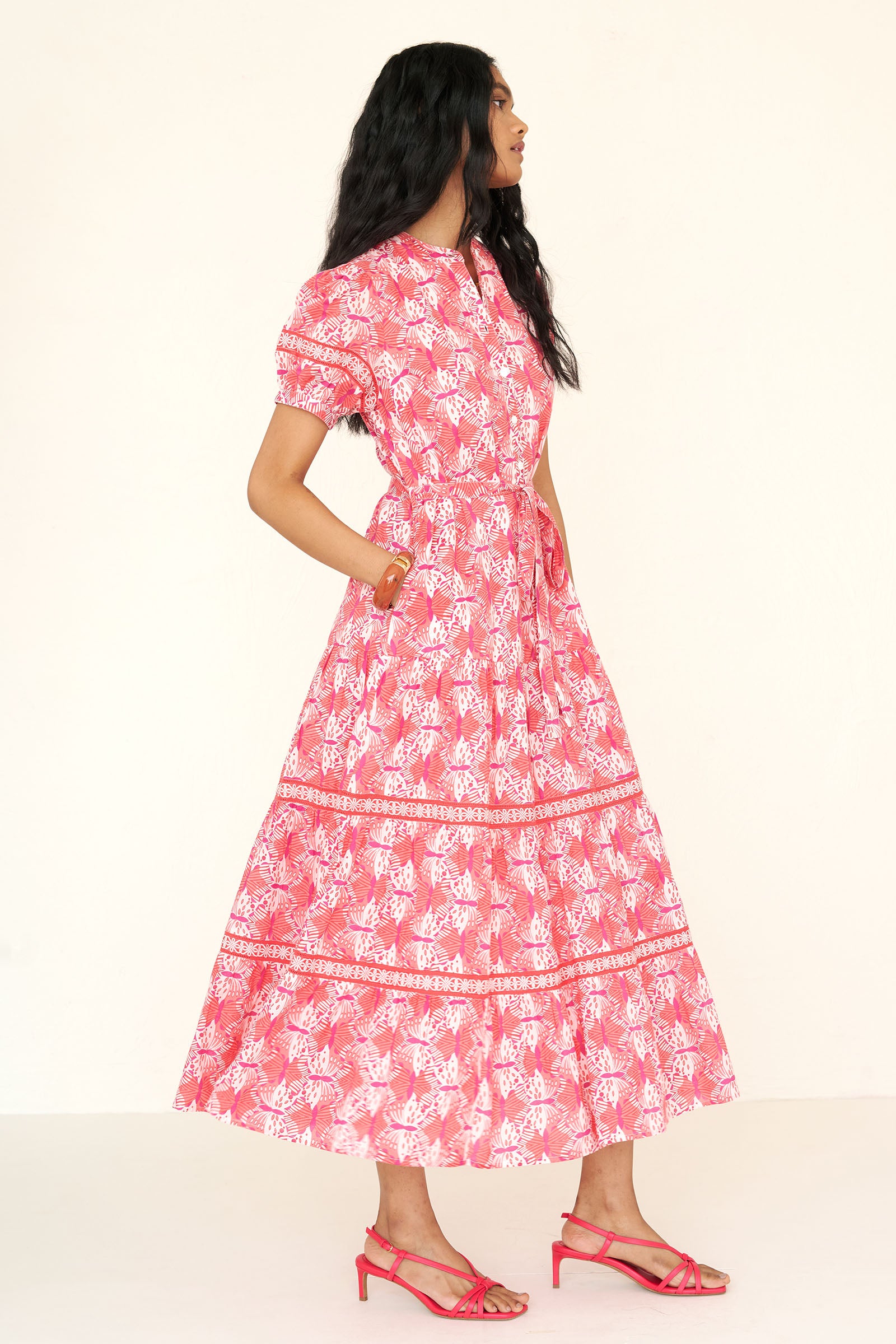 Side view of a woman standing wearing the Saylor dress in Flutter print with her hand in pocket. It features a stand collar, tiered skirt, detachable belt, and celtic-knot ladder lace detailing in a full-length shirtdress silhouette.