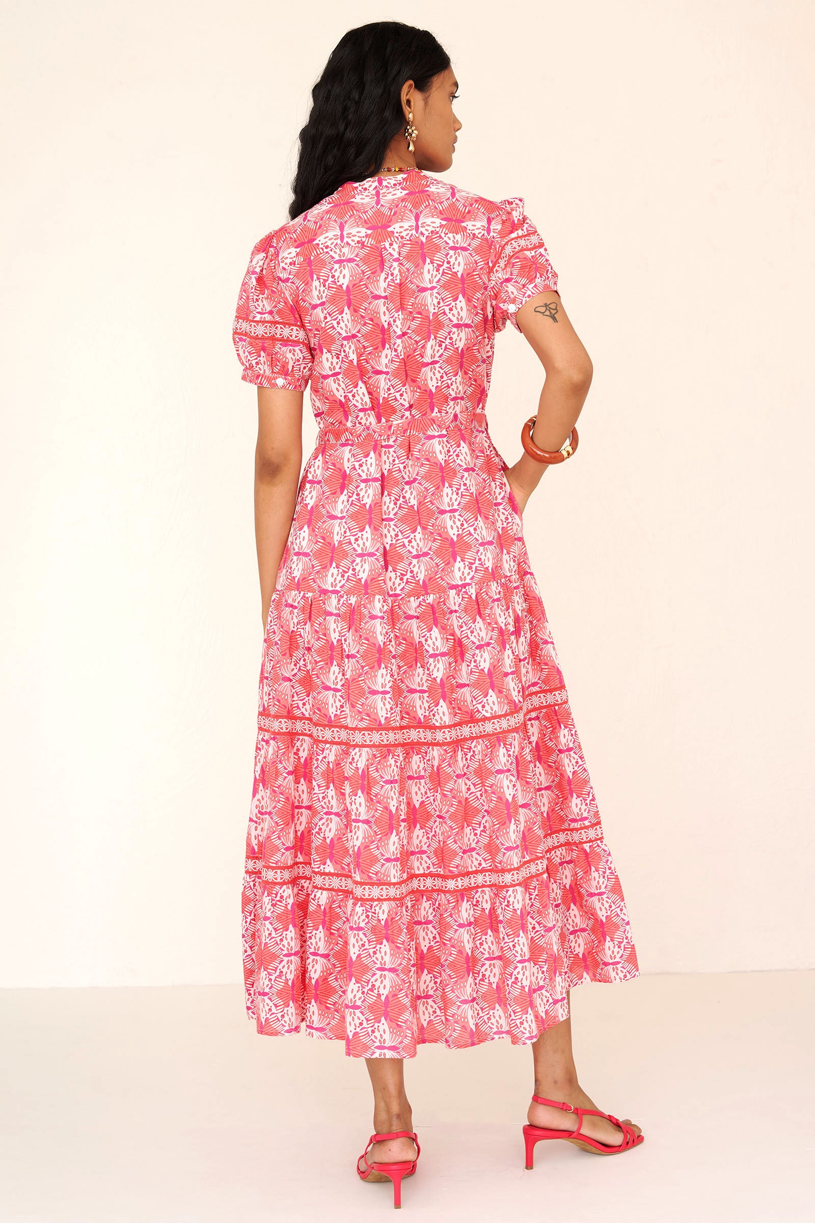 Back view of a woman standing wearing the Saylor dress in Flutter print. It features a stand collar, tiered skirt, detachable belt, and celtic-knot ladder lace detailing in a full-length shirtdress silhouette.