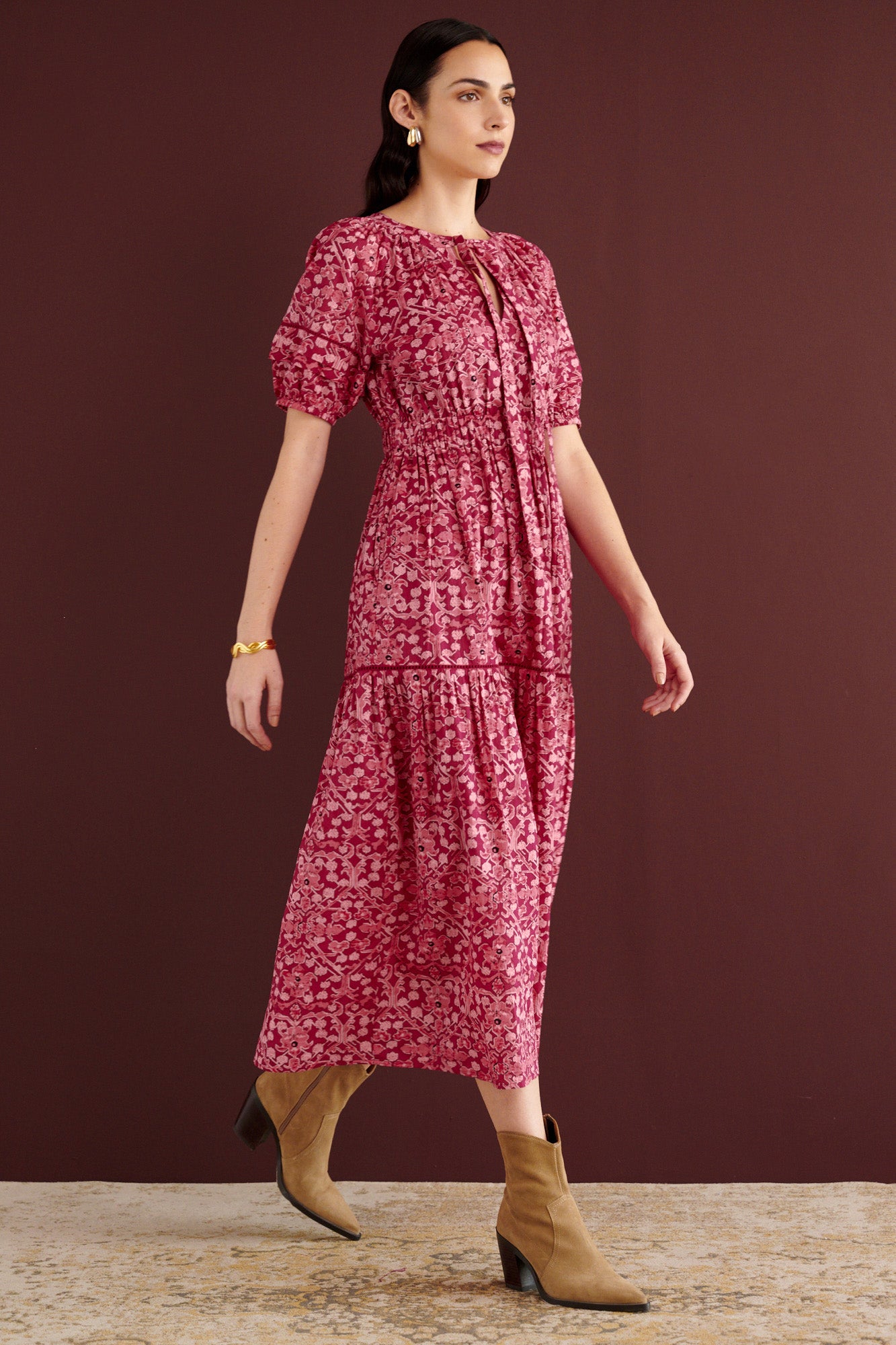 Side view of woman walking sideways. She is wearing the Selma full-length V-neck dress in Ottoman print, with tie-neck, elastic waist, lace insets at skirt and sleeves, smooth cotton cambric, side-seam pockets, and fully lined construction.