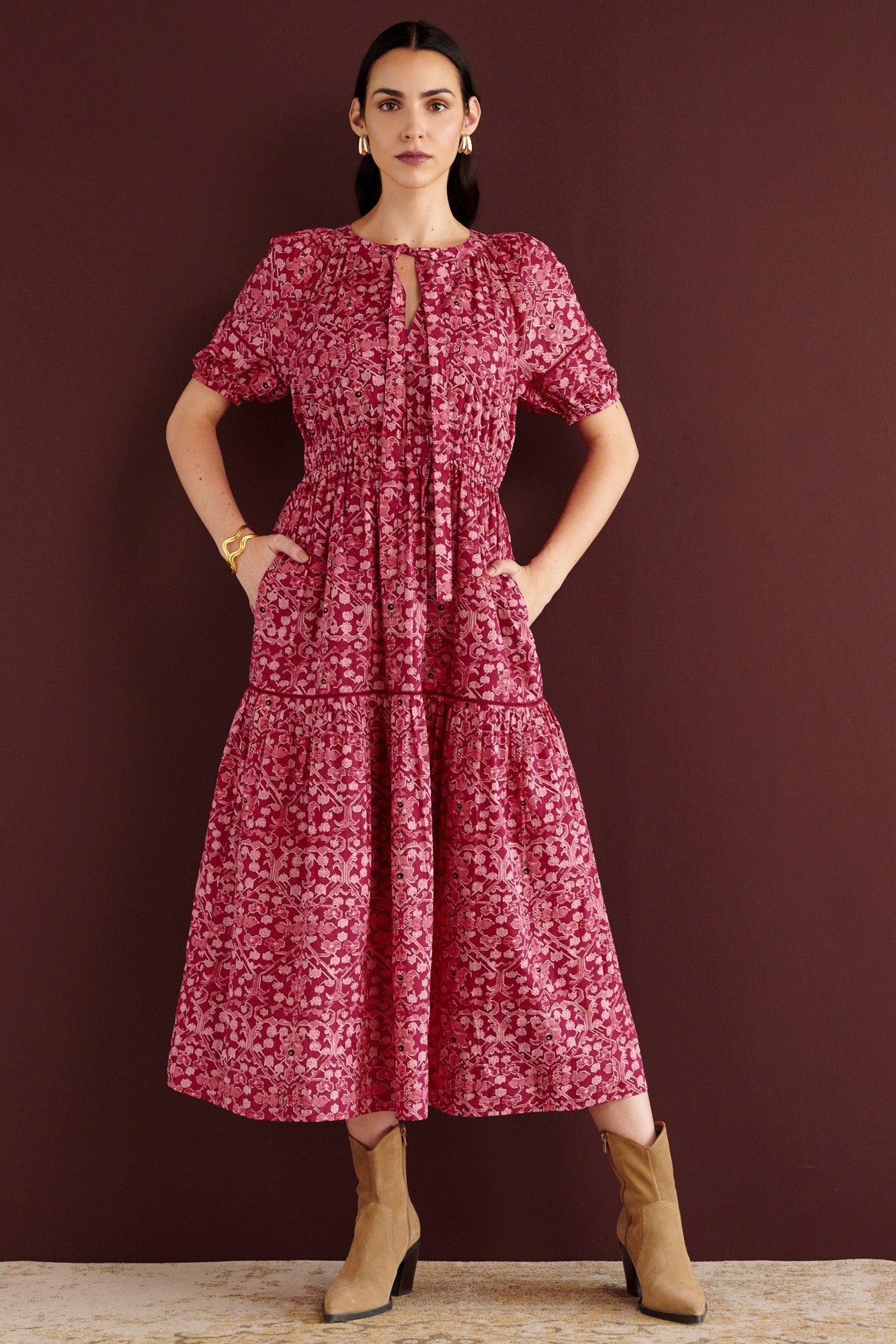 Front view of woman standing with hands in pocket. She is wearing the Selma full-length V-neck dress in Ottoman print, with tie-neck, elastic waist, lace insets at skirt and sleeves, smooth cotton cambric, side-seam pockets, and fully lined construction.