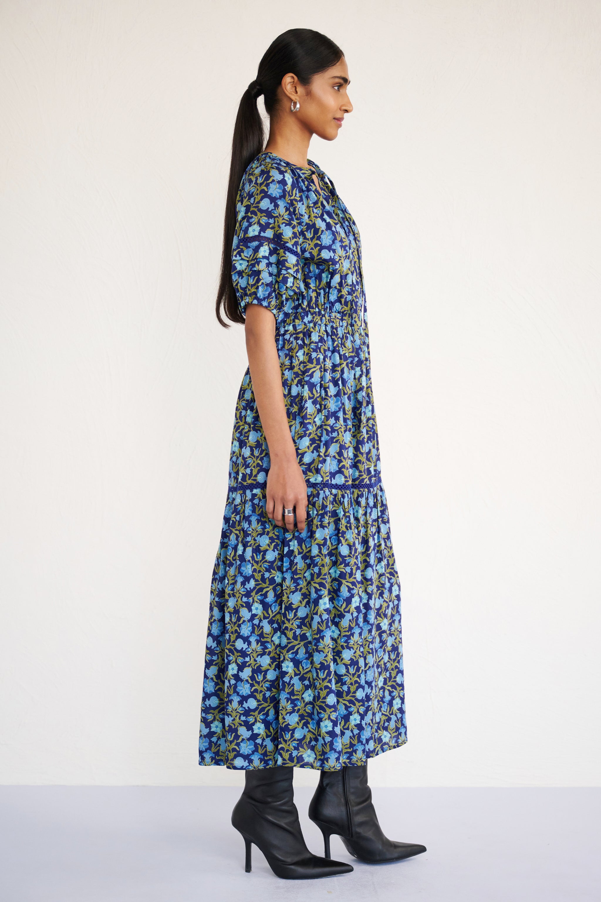 Side view of woman wearing a full-length V-neck dress with tie-neck detail, lace insets on the skirt and sleeves, and a flowing pomegranate vine print.