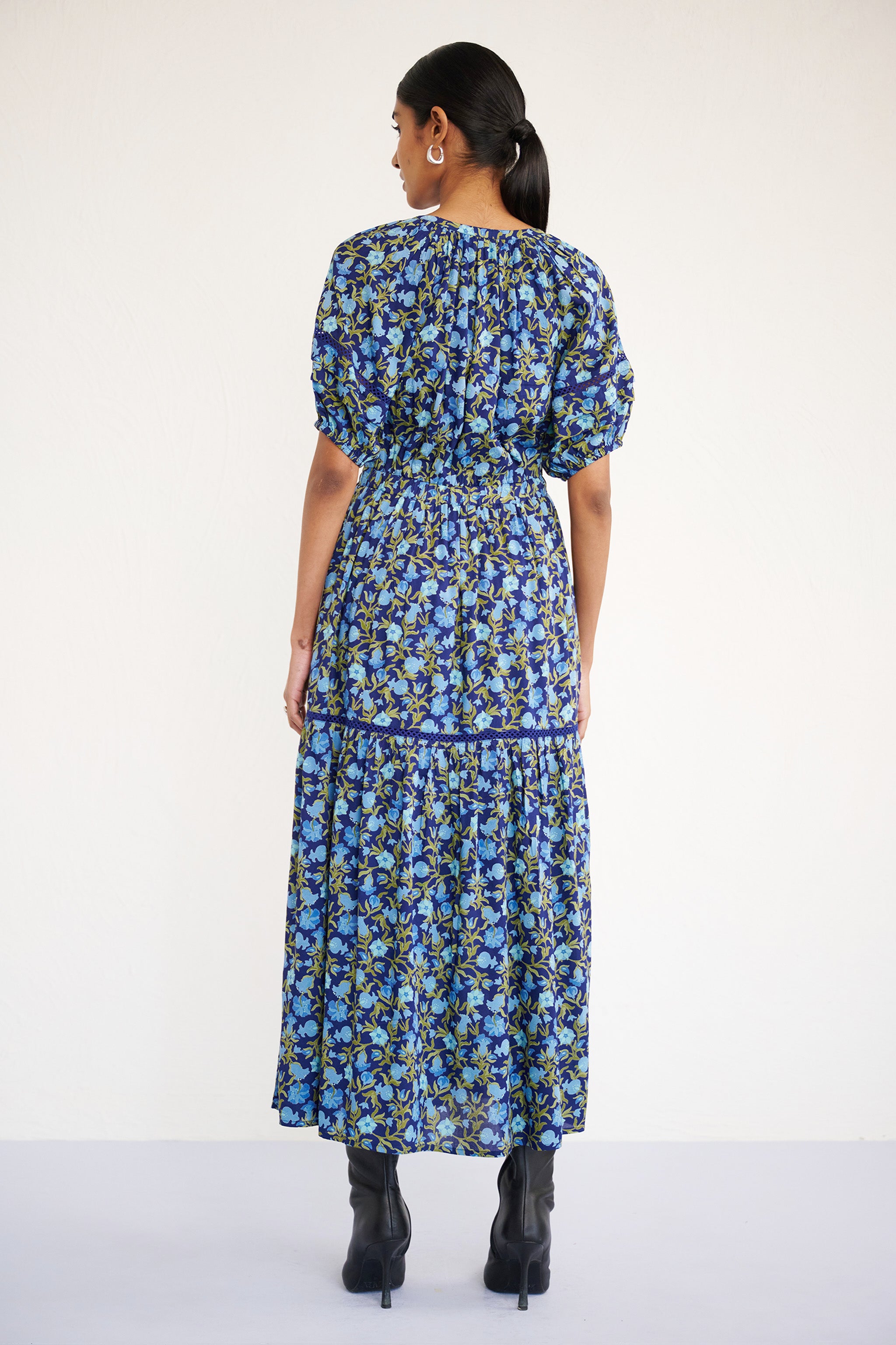 Back view of woman wearing a full-length V-neck dress with tie-neck detail, lace insets on the skirt and sleeves, and a flowing pomegranate vine print.