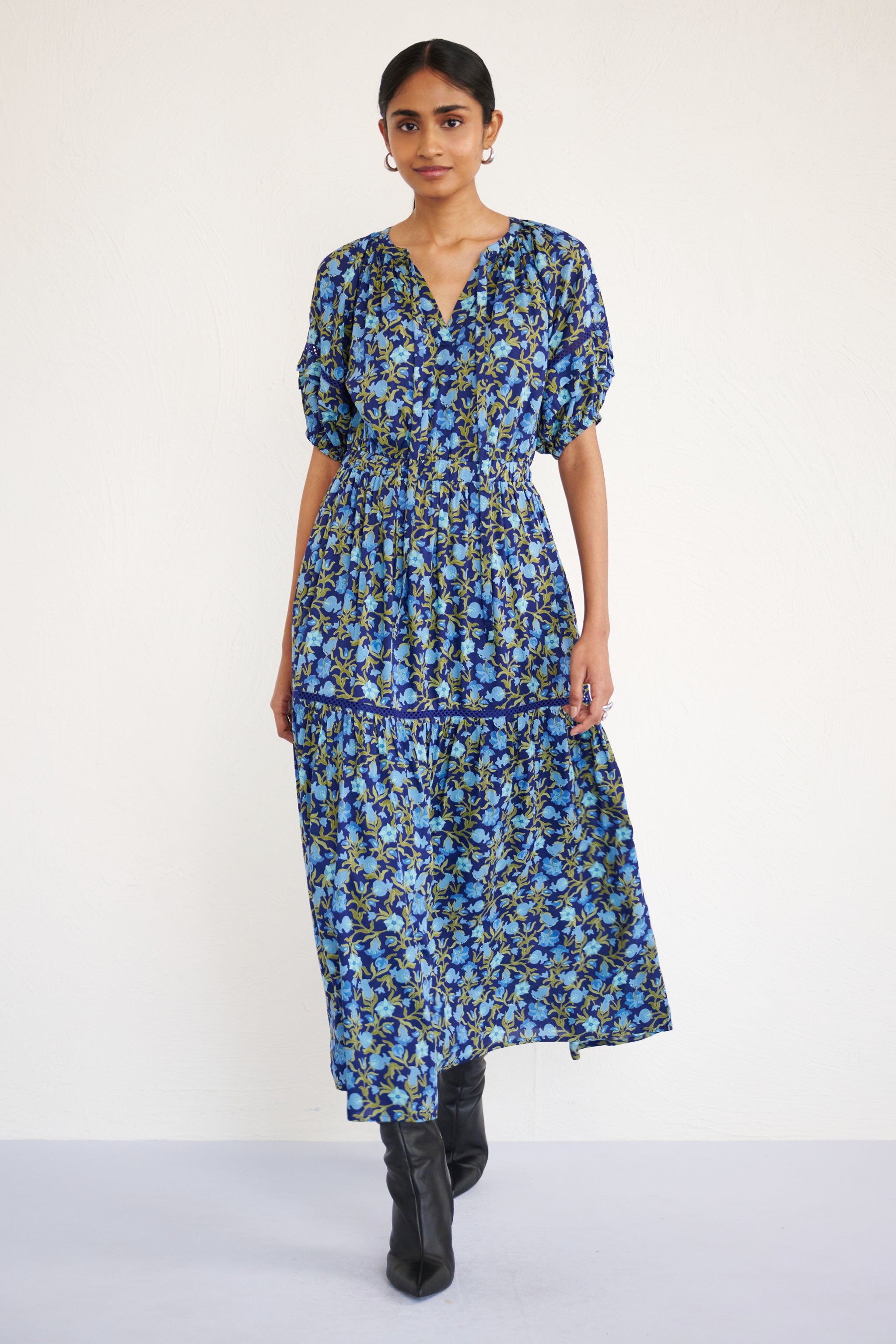 Front view of woman wearing a full-length V-neck dress with tie-neck detail, lace insets on the skirt and sleeves, and a flowing pomegranate vine print.