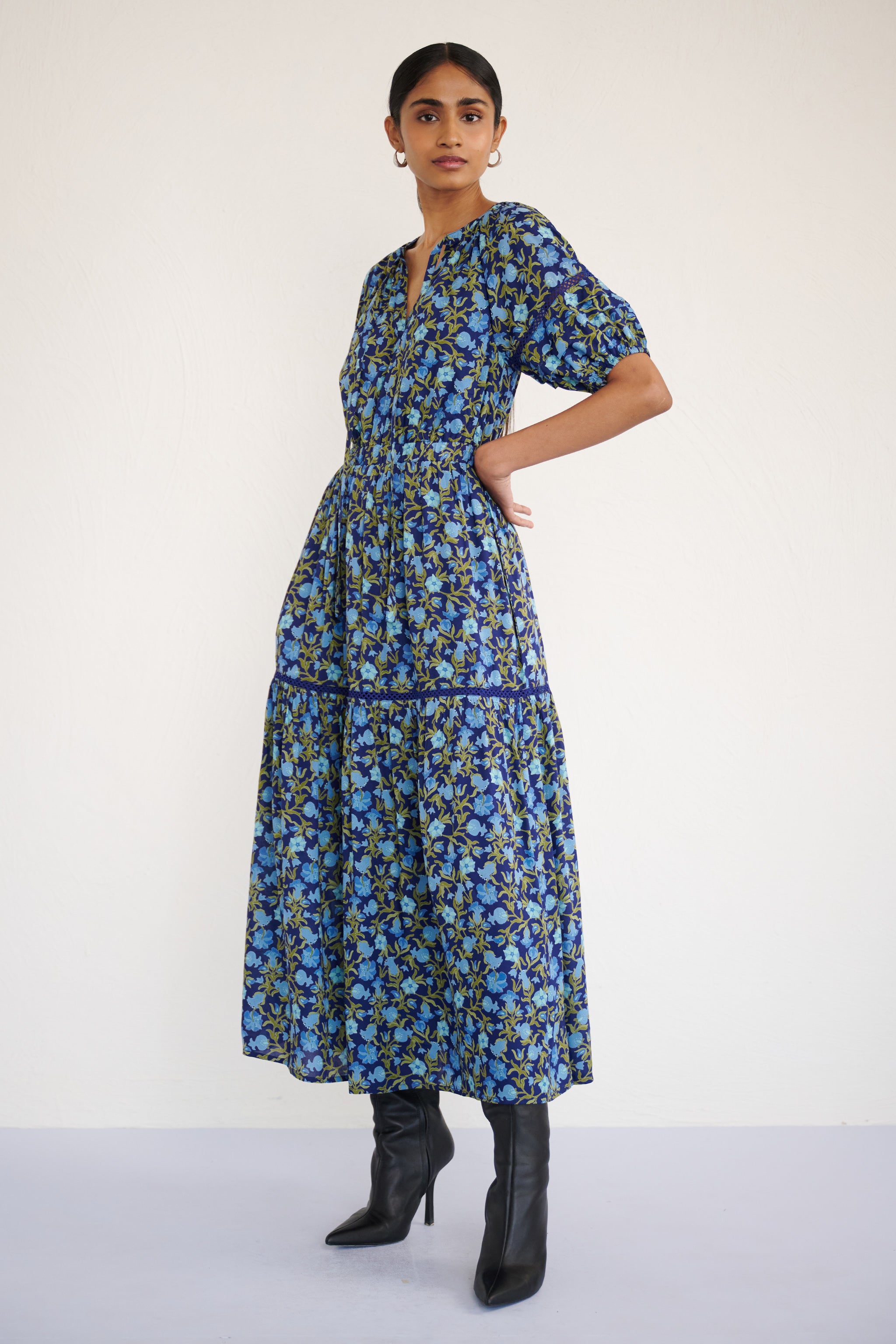 Front view of woman wearing a full-length V-neck dress with tie-neck detail, lace insets on the skirt and sleeves, and a flowing pomegranate vine print.
