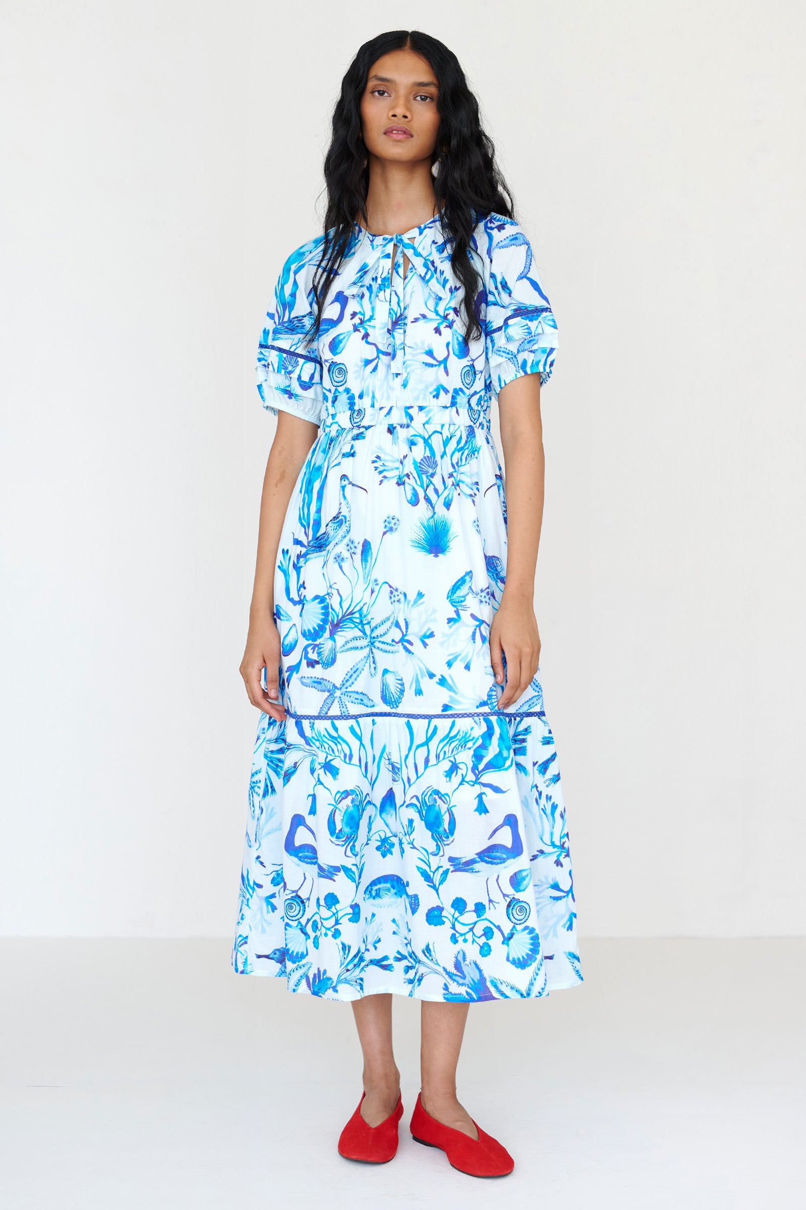 Front view of a woman standing wearing the Selma dress in Tidepool print, featuring a V-neck with tie, elastic waist, ladder lace insets, full-length skirt and side-seam pockets.