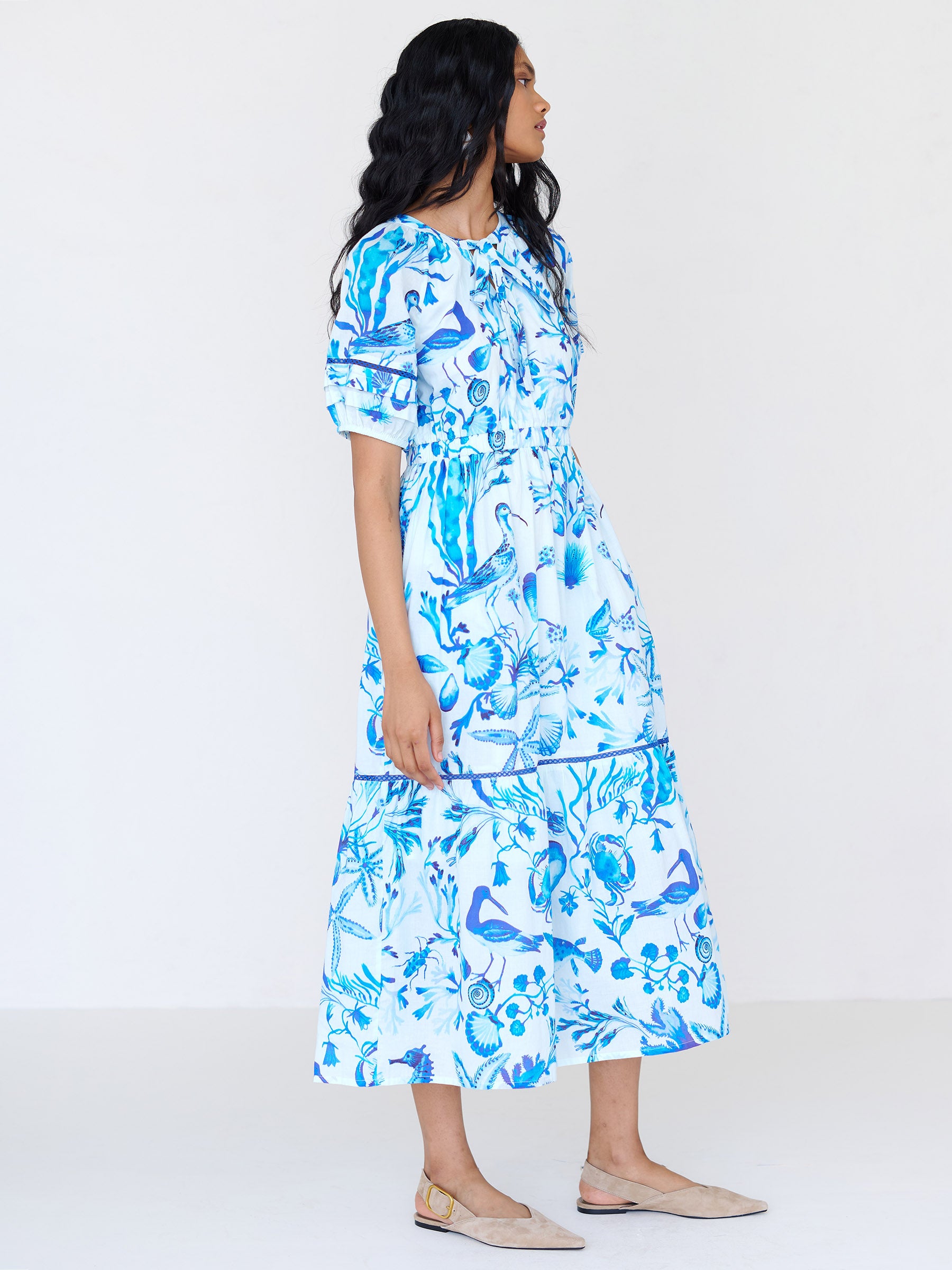 Side view of a woman standing. She is wearing the Selma full-length V-neck dress in tonal Tidepool print. It features a tie-neck, elasticated waist, ladder lace detailing, and side-seam pockets.