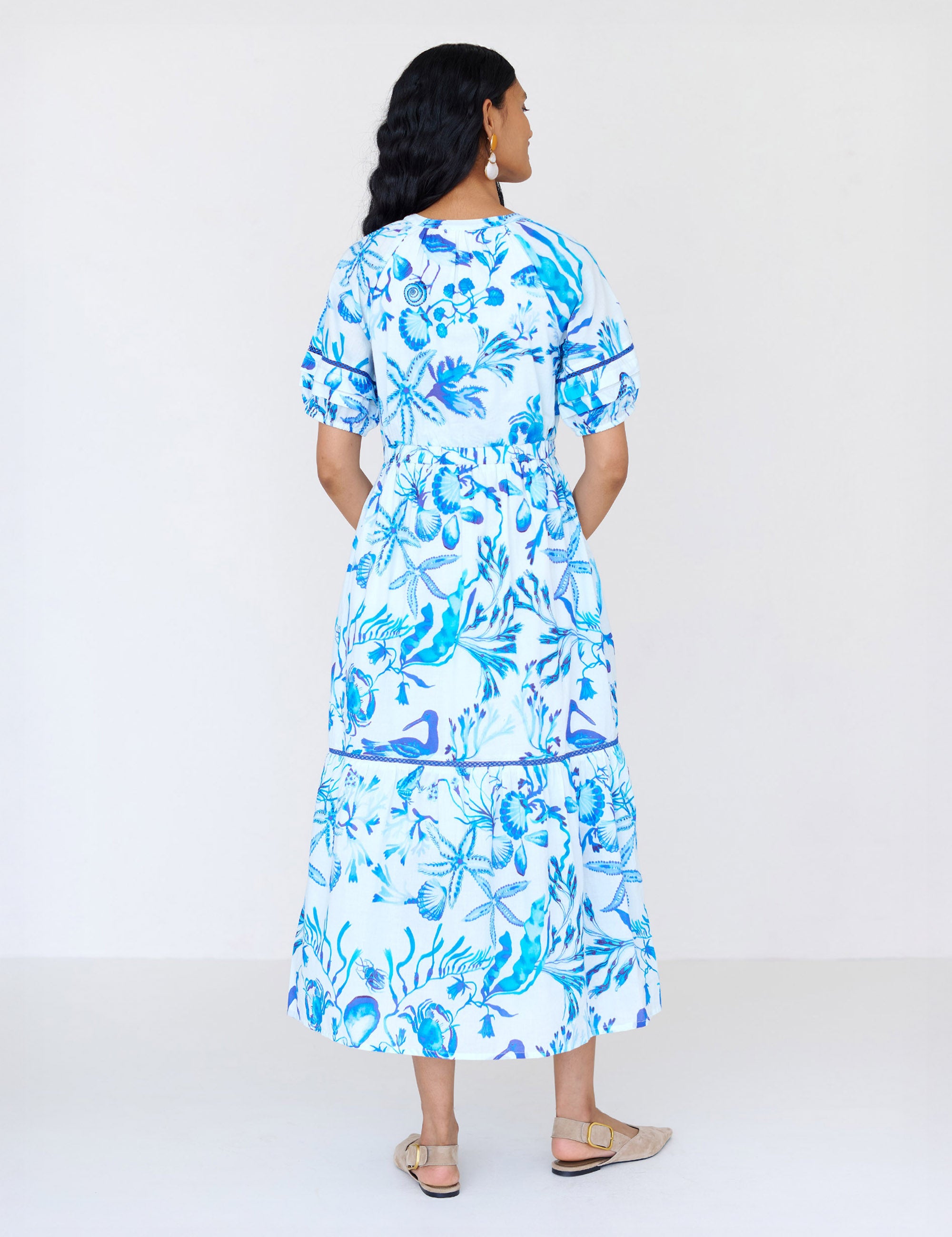 Back view of a woman standing. She is wearing the Selma full-length V-neck dress in tonal Tidepool print. It features a tie-neck, elasticated waist, ladder lace detailing, and side-seam pockets.