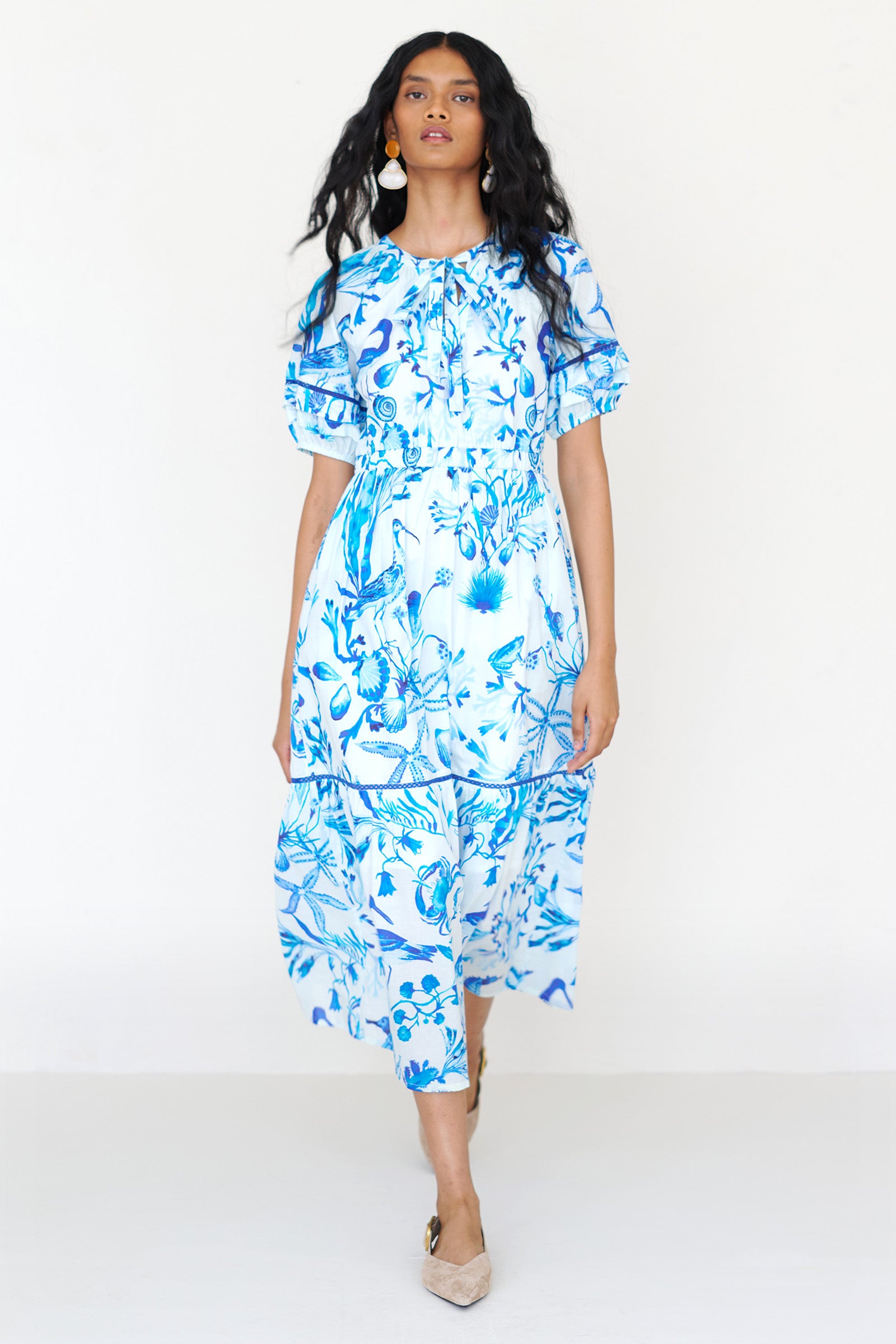 Front view of a woman walking wearing the Selma dress in Tidepool print, featuring a V-neck with tie, elastic waist, ladder lace insets, full-length skirt and side-seam pockets.