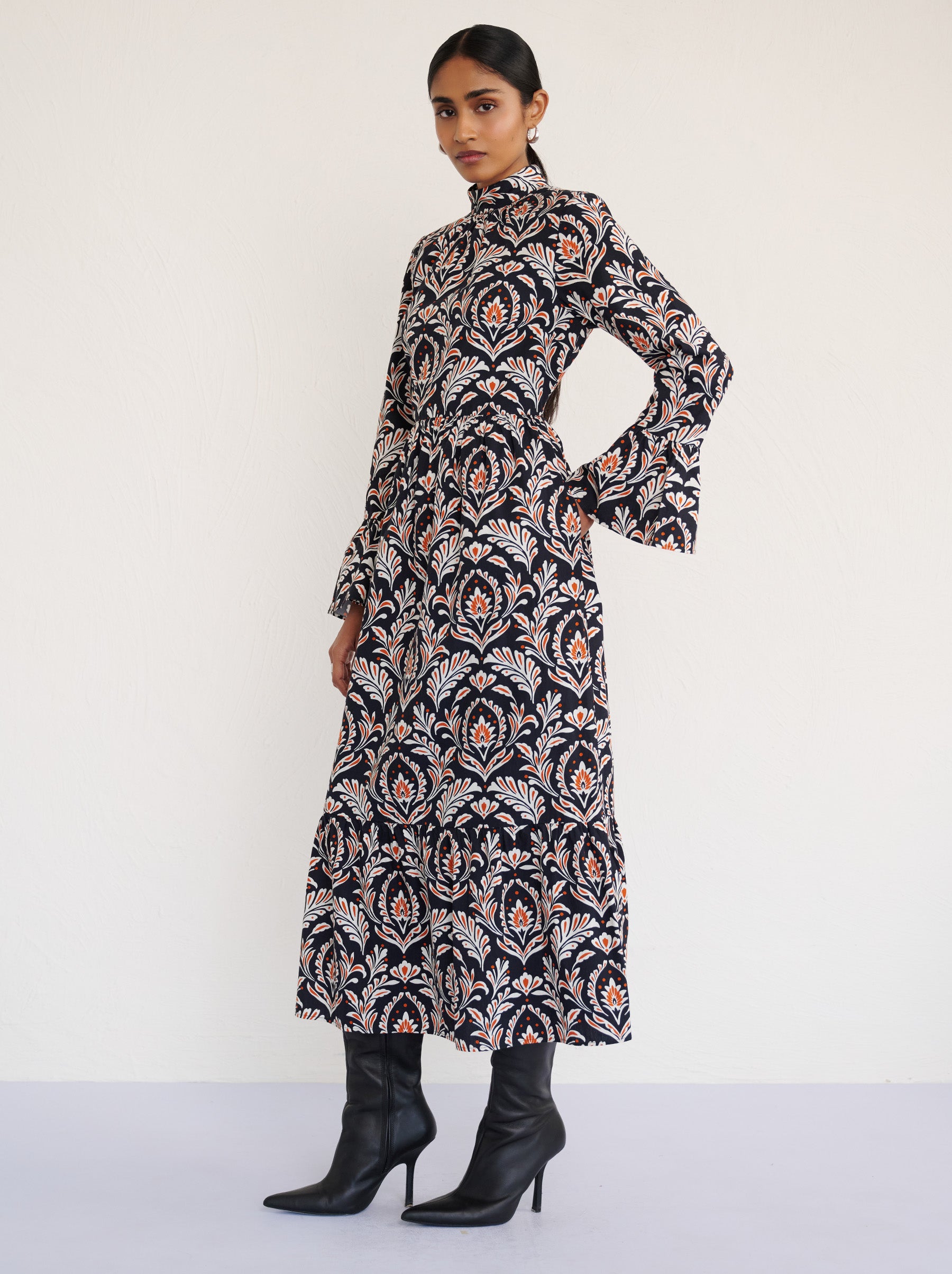 Side view of a model standing with right hand in pocket. She is wearing the Sumi long-sleeve dress in crisp cotton poplin in Akbar Galaxy print. It is featuring a high neckline, ruffled sleeves, a flowing skirt, and side-seam pockets.