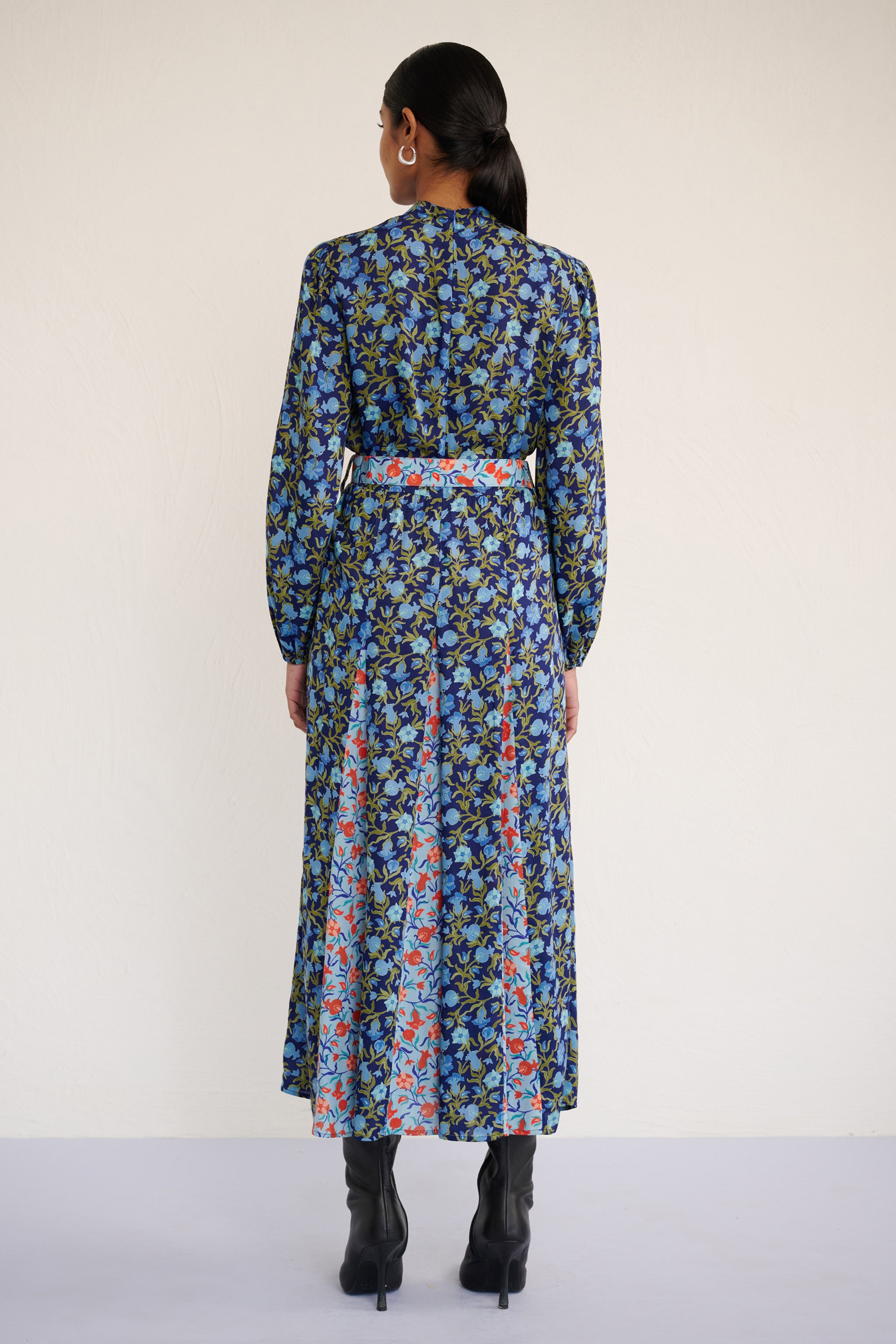 Back view of woman wearing a full-length dress with a stand collar, fitted waist belt, and flowing skirt featuring contrast print inserts and a pomegranate vine motif.
