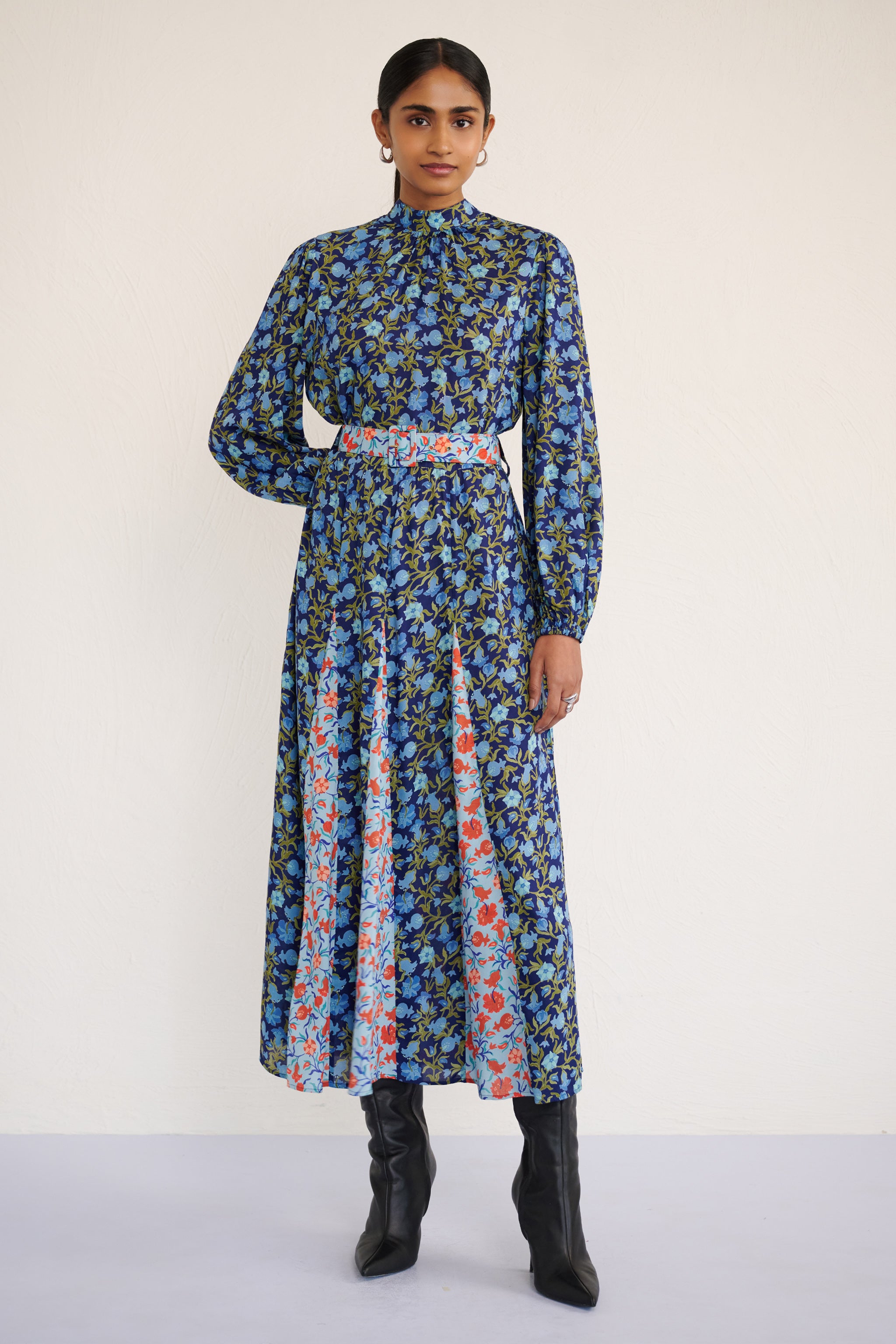 Front view of woman wearing a full-length dress with a stand collar, fitted waist belt, and flowing skirt featuring contrast print inserts and a pomegranate vine motif.