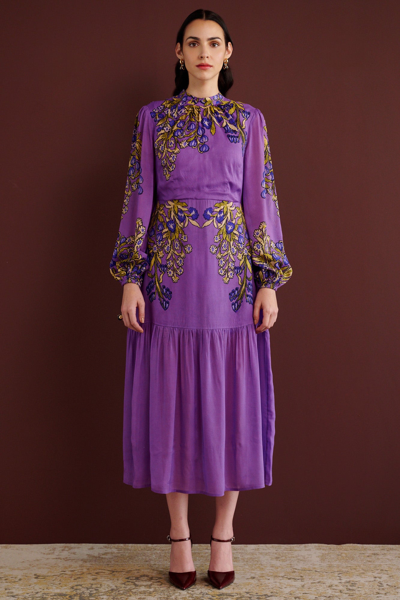Front view of woman standing. She is wearing the Sumina dress in the Unicorn Tapestry print. It is a purple viscose voile midi dress with a fitted crew neck, and tiered skirt, featuring intricate floral and mythical motifs inspired by 15th-century Scottish embroidery.