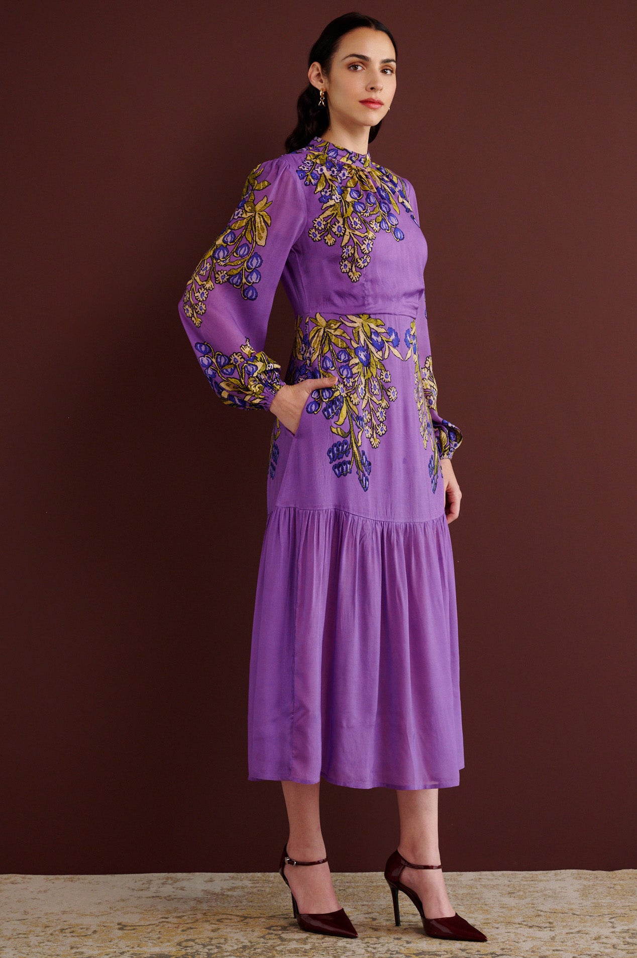 Side view of woman standing with hand in pocket. She is wearing the Sumina dress in the Unicorn Tapestry print. It is a purple viscose voile midi dress with a fitted crew neck, and tiered skirt, featuring intricate floral and mythical motifs inspired by 15th-century Scottish embroidery.