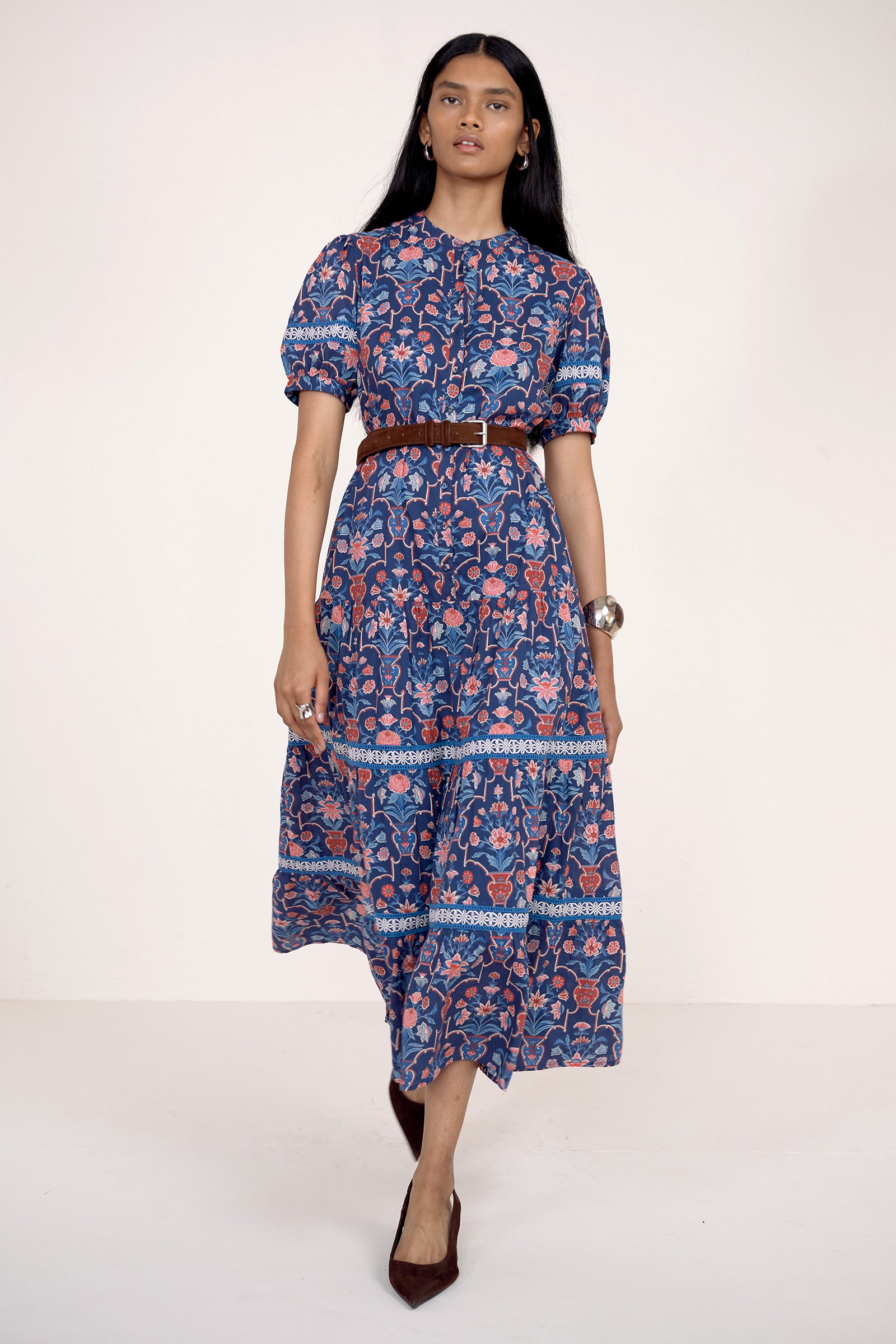 Front view of model stepping forward. She is wearing the Saylor dress in Floral Vase print. It features a shirtdress silhouette, lace trim, tiered skirt, and detachable waist belt.