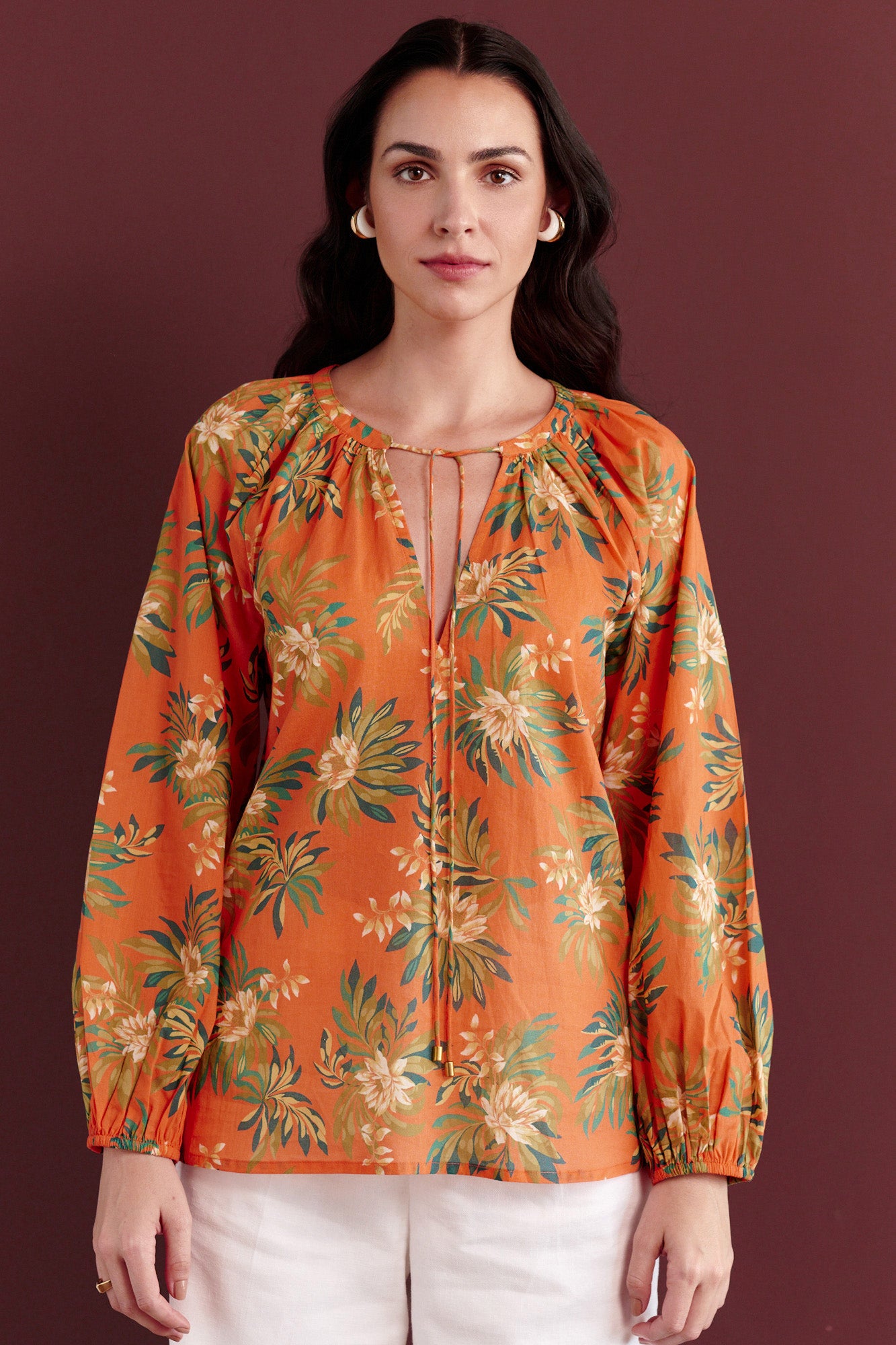 Front view of woman standing. She is wearing the Tara crew-neck shirt in the Artichoke Flower print paired with white trouser. It is a breezy cotton voile top with a tie-neck detail and a relaxed, tropical-inspired design.