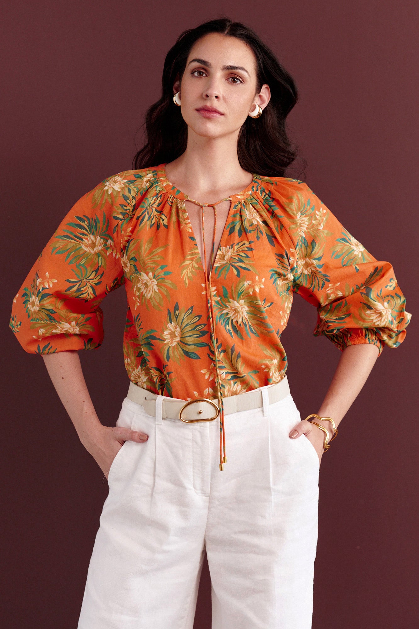 Front view of woman standing. She is wearing the Tara crew-neck shirt in the Artichoke Flower print paired with white trouser. It is a breezy cotton voile top with a tie-neck detail and a relaxed, tropical-inspired design.