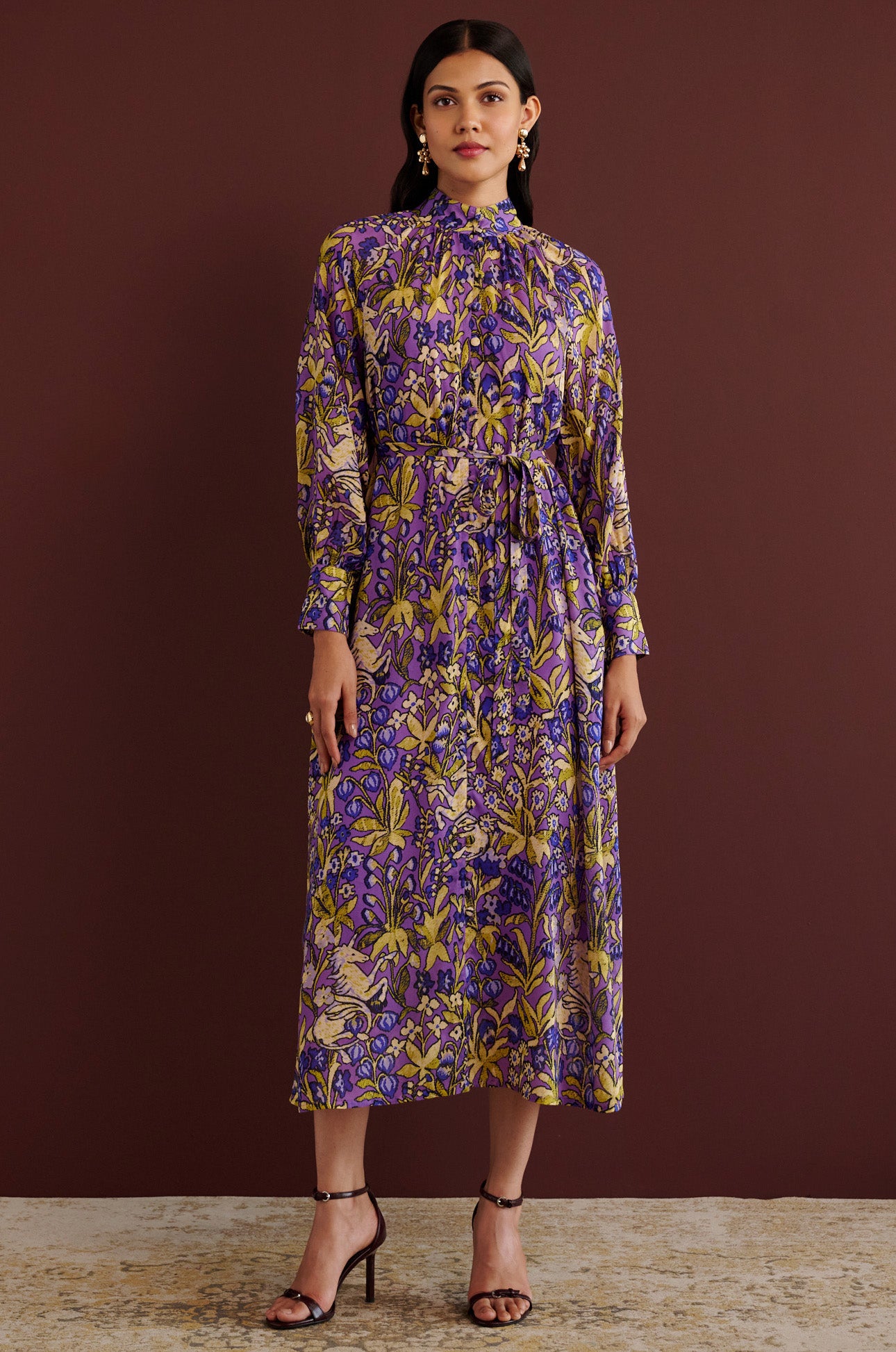 Front view of woman standing. She is wearing the Tara dress in the Unicorn Tapestry print. It is a flowing purple viscose voile dress with a stand collar, gathered neckline and sleeves, fabric-covered buttons, and a removable belt, featuring intricate floral and mythical motifs inspired by the Scottish Highlands.