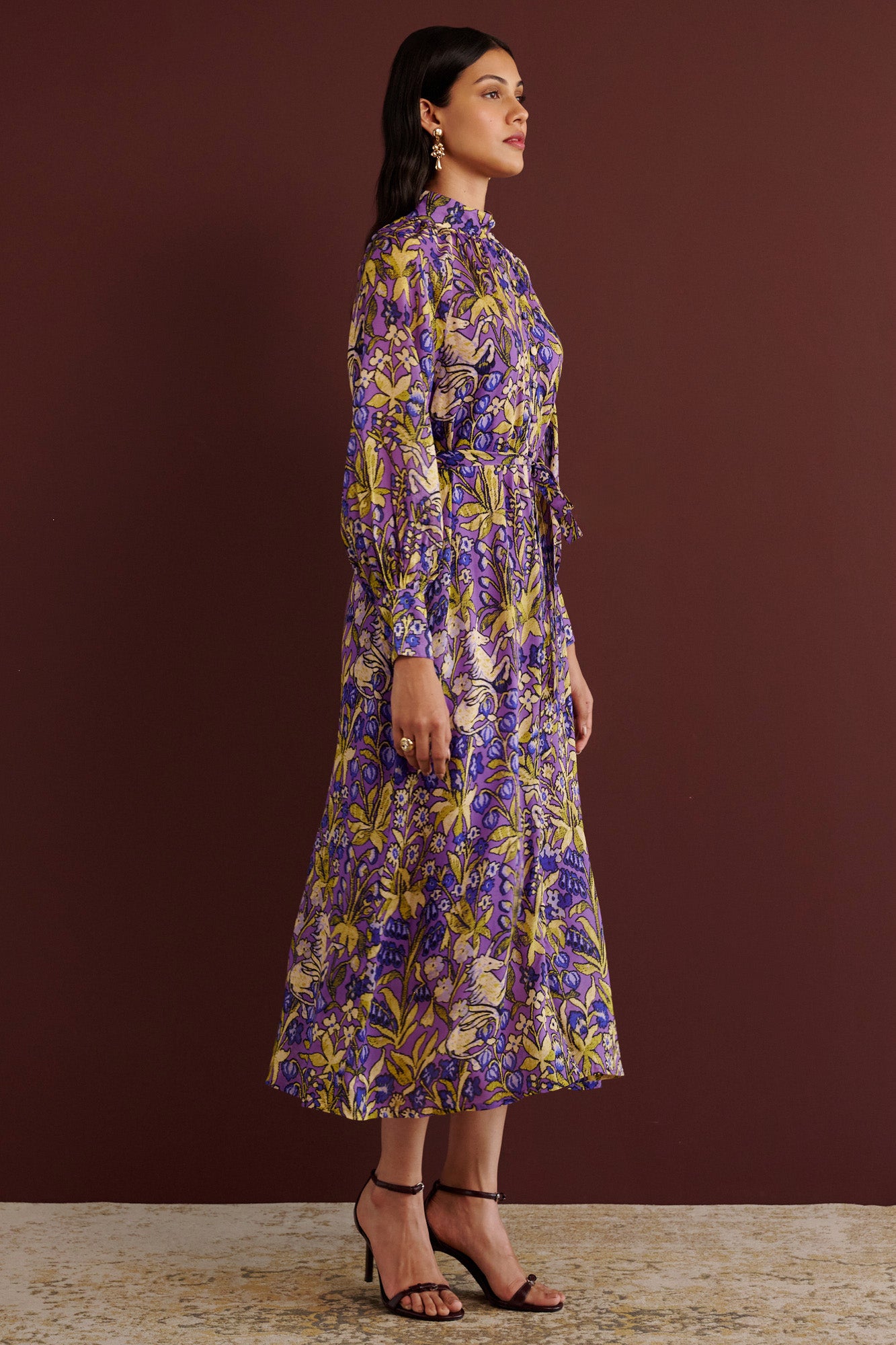 Side view of woman standing. She is wearing the Tara dress in the Unicorn Tapestry print. It is a flowing purple viscose voile dress with a stand collar, gathered neckline and sleeves, fabric-covered buttons, and a removable belt, featuring intricate floral and mythical motifs inspired by the Scottish Highlands.