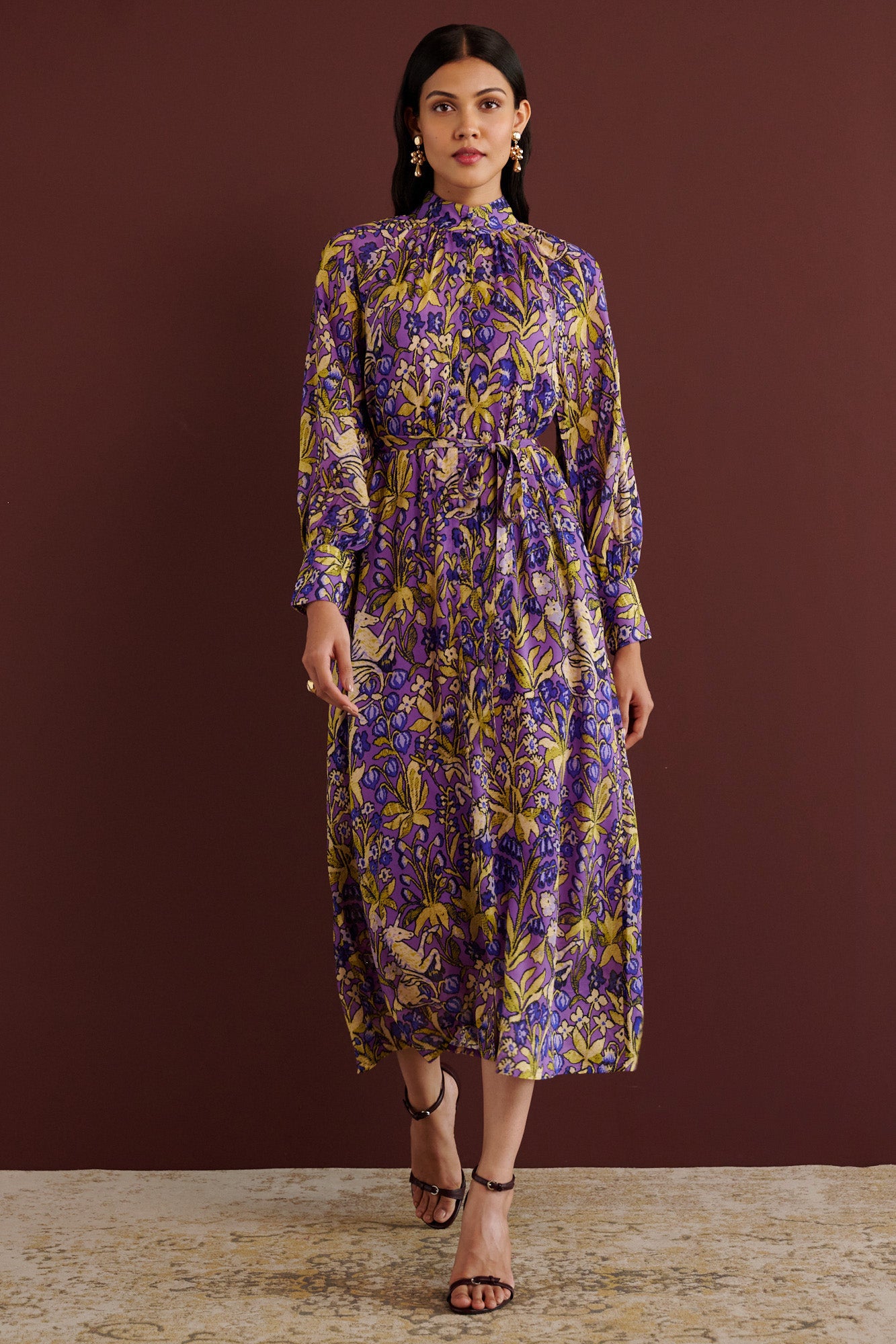 Front view of woman walking. She is wearing the Tara dress in the Unicorn Tapestry print. It is a flowing purple viscose voile dress with a stand collar, gathered neckline and sleeves, fabric-covered buttons, and a removable belt, featuring intricate floral and mythical motifs inspired by the Scottish Highlands.