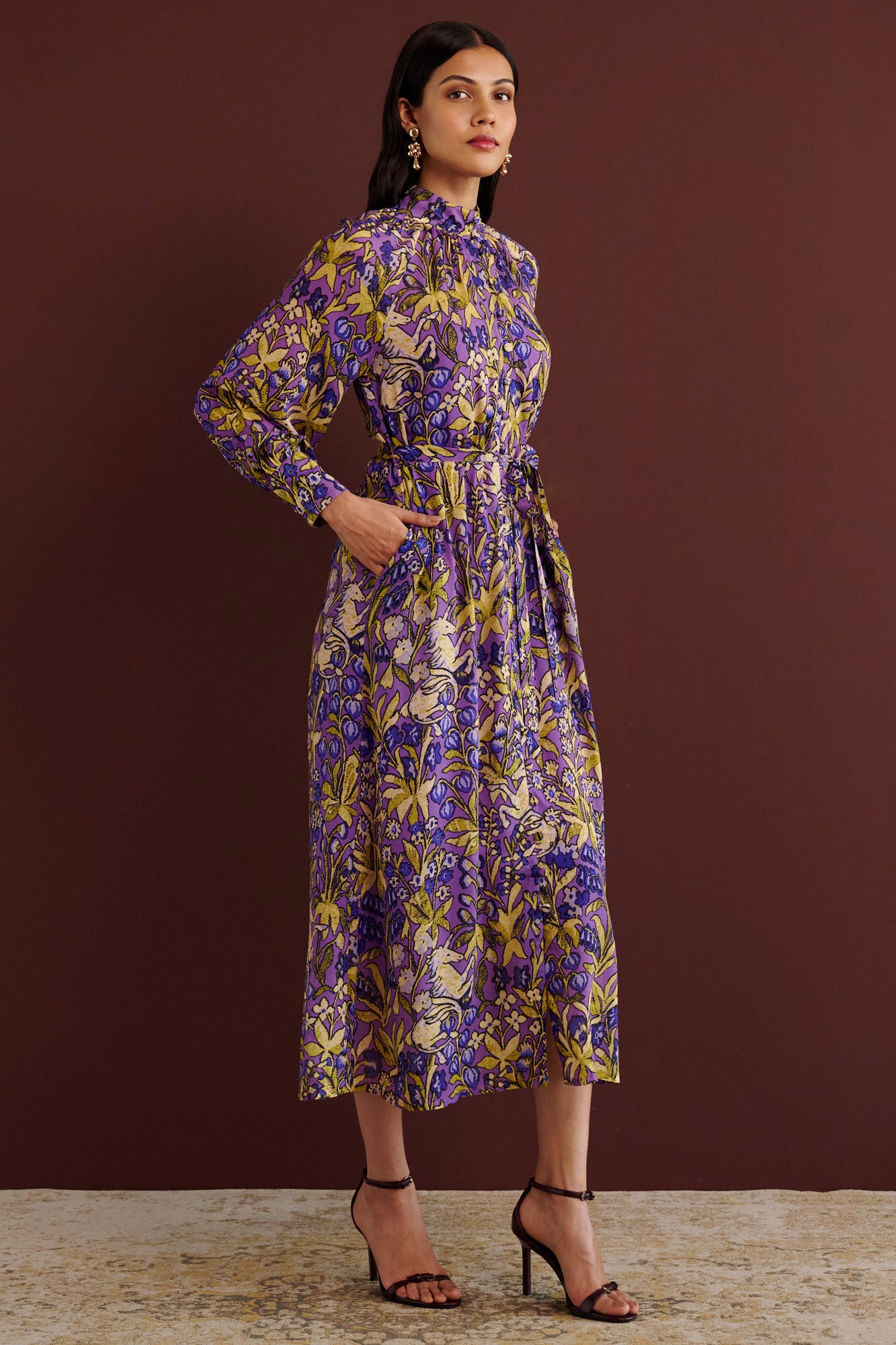 Side view of woman standing with hands in pocket. She is wearing the Tara dress in the Unicorn Tapestry print. It is a flowing purple viscose voile dress with a stand collar, gathered neckline and sleeves, fabric-covered buttons, and a removable belt, featuring intricate floral and mythical motifs inspired by the Scottish Highlands.