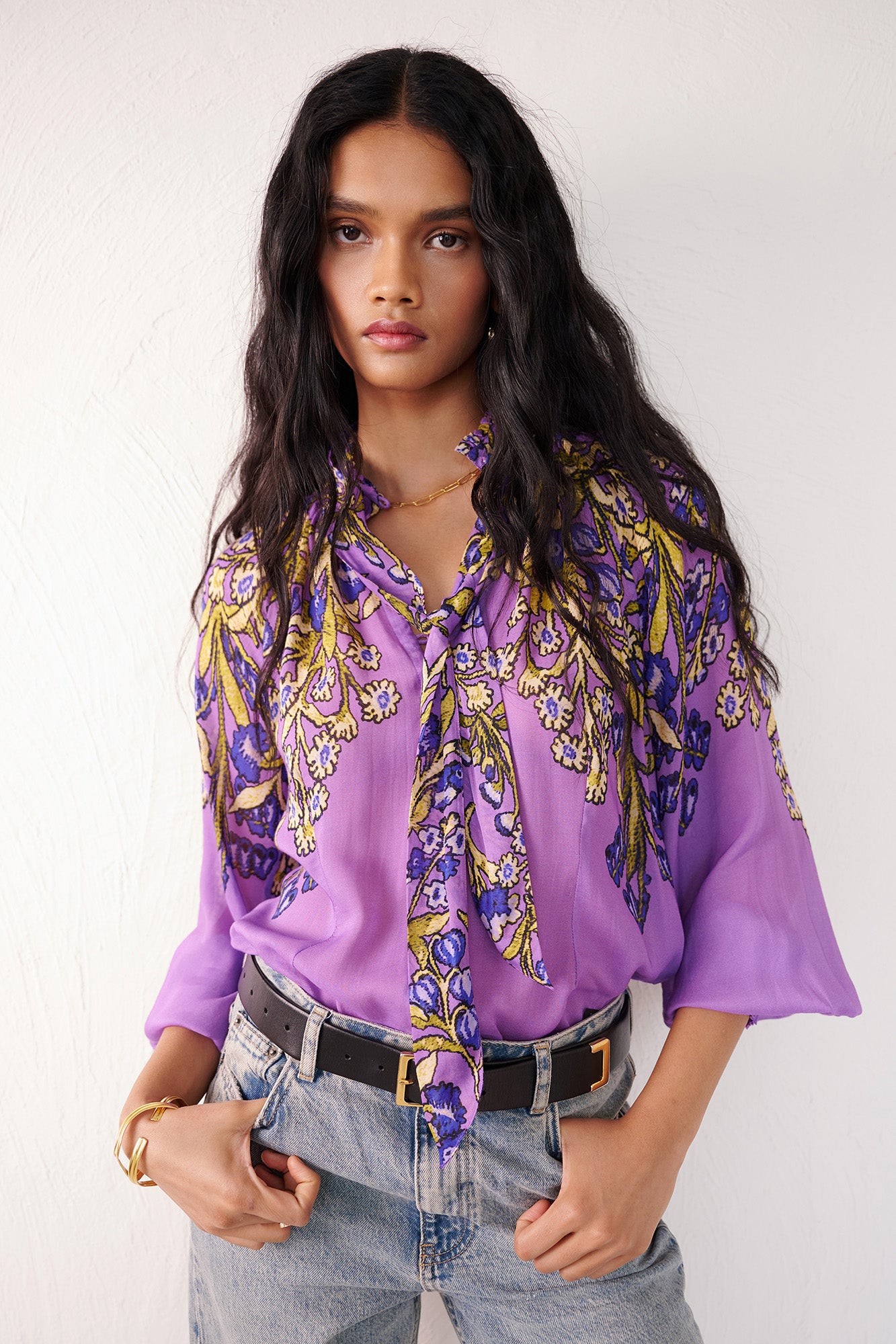 Front view of woman standing. She is wearing the Tara tie-neck shirt in the Unicorn Tapestry print, a purple viscose voile blouse with yellow and blue floral motifs. It features high ruffled stand collar with a removable tie at the neck, and gathered sleeves, paired with denim pants and a black belt.
