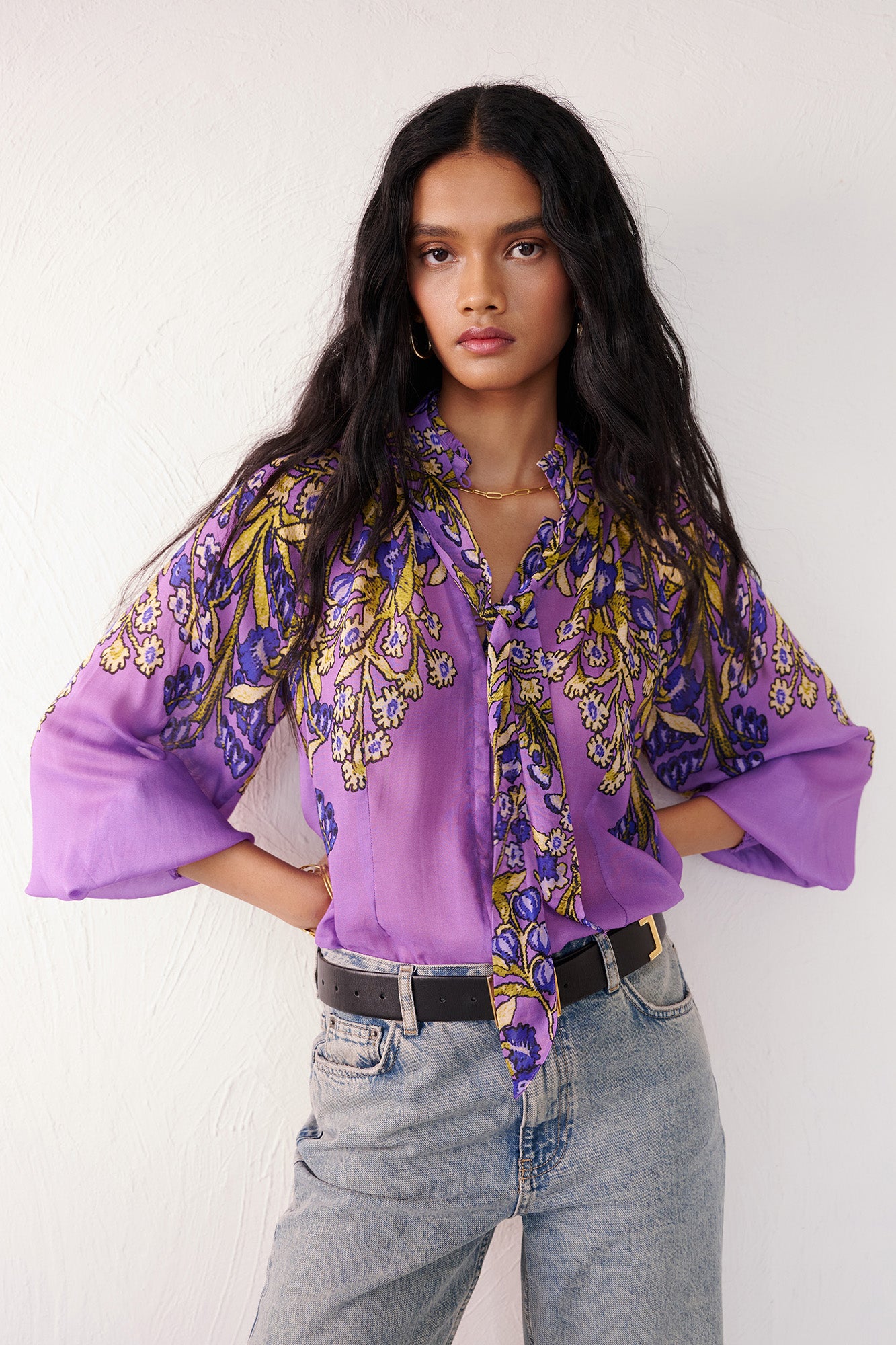 Front view of woman standing. She is wearing the Tara tie-neck shirt in the Unicorn Tapestry print, a purple viscose voile blouse with yellow and blue floral motifs. It features high ruffled stand collar with a removable tie at the neck, and gathered sleeves, paired with denim pants and a black belt.