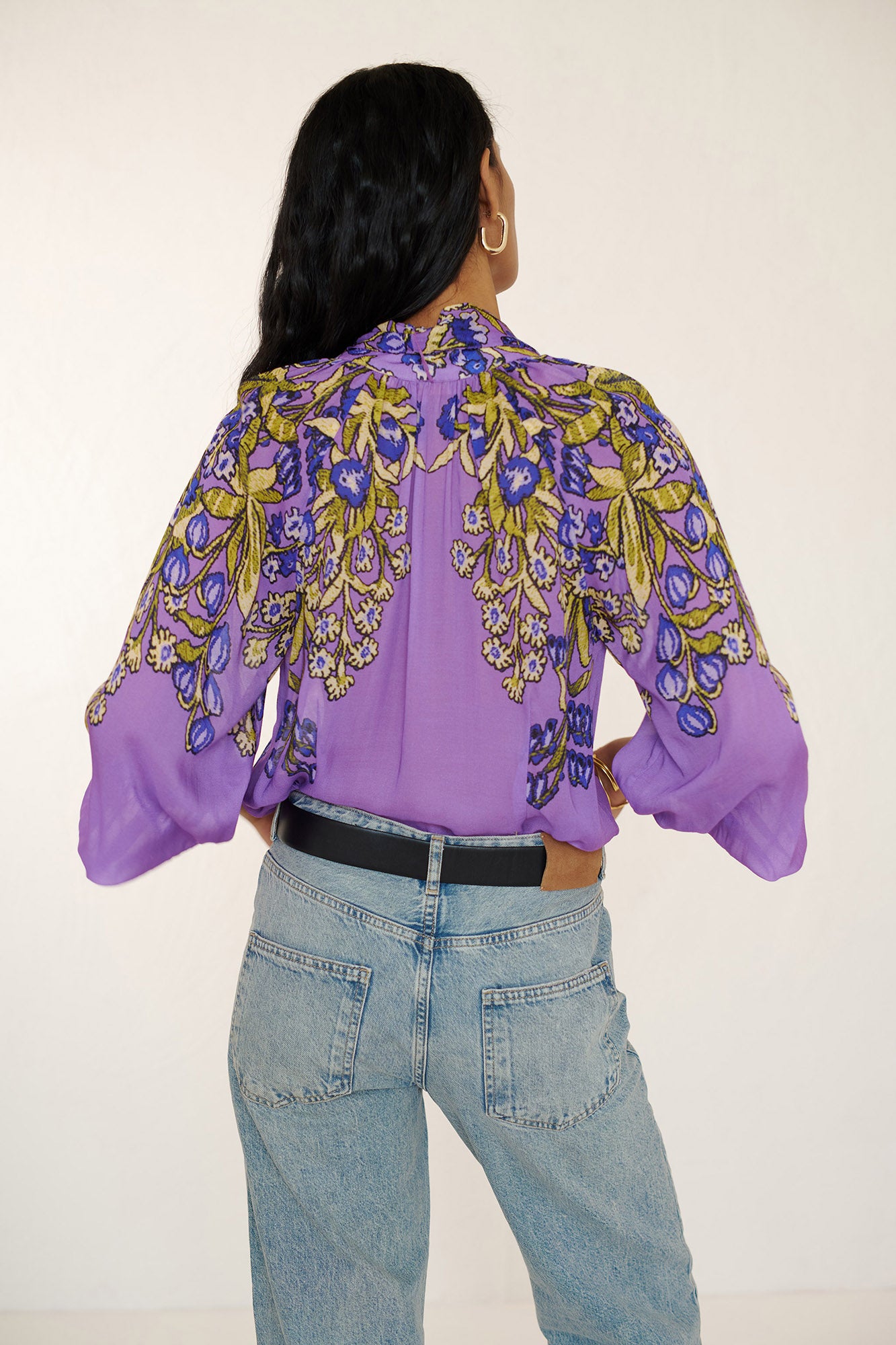 Back view of woman standing. She is wearing the Tara tie-neck shirt in the Unicorn Tapestry print, a purple viscose voile blouse with yellow and blue floral motifs. It features high ruffled stand collar with a removable tie at the neck, and gathered sleeves, paired with denim pants and a black belt.