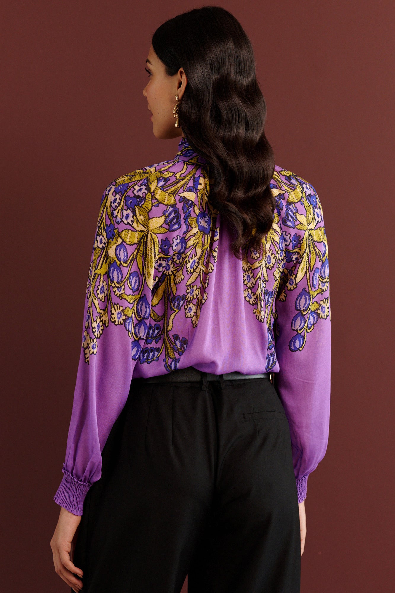 Back view of woman standing. She is wearing the Tara tie-neck shirt in the Unicorn Tapestry print, a purple viscose voile blouse with yellow and blue floral motifs, high ruffled collar, and gathered sleeves, paired with black pleated trousers and a black belt.