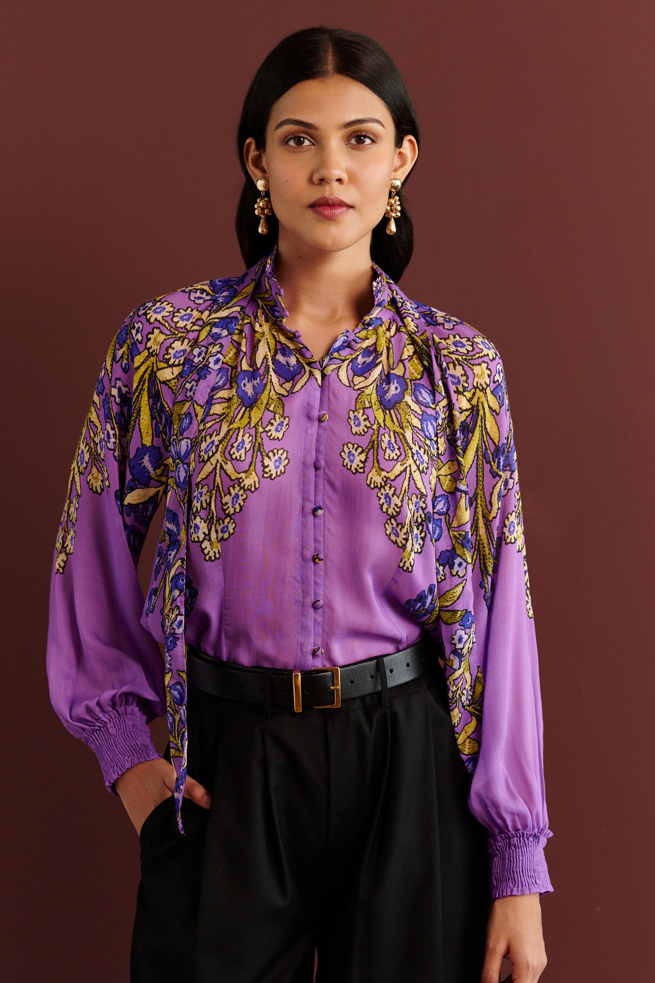 Front view of woman standing. She is wearing the Tara tie-neck shirt in the Unicorn Tapestry print, a purple viscose voile blouse with yellow and blue floral motifs, high ruffled collar, and gathered sleeves, paired with black pleated trousers and a black belt.