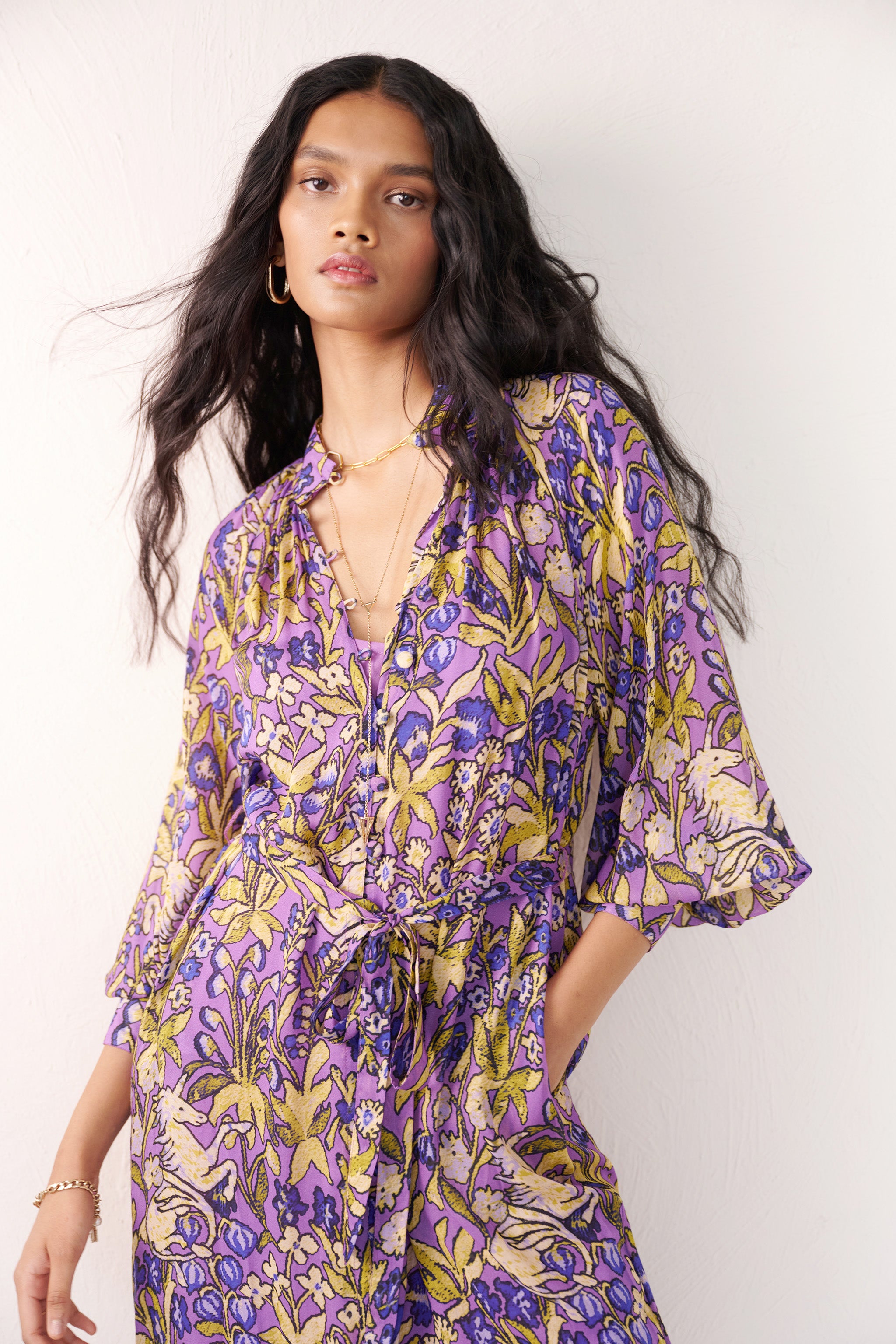 Front view of close up of woman standing with a hand in pocket. She is wearing the Tara dress in the Unicorn Tapestry print. It is a flowing purple viscose voile dress with a stand collar, gathered neckline and sleeves, fabric-covered buttons, and a removable belt, featuring intricate floral and mythical motifs inspired by the Scottish Highlands.