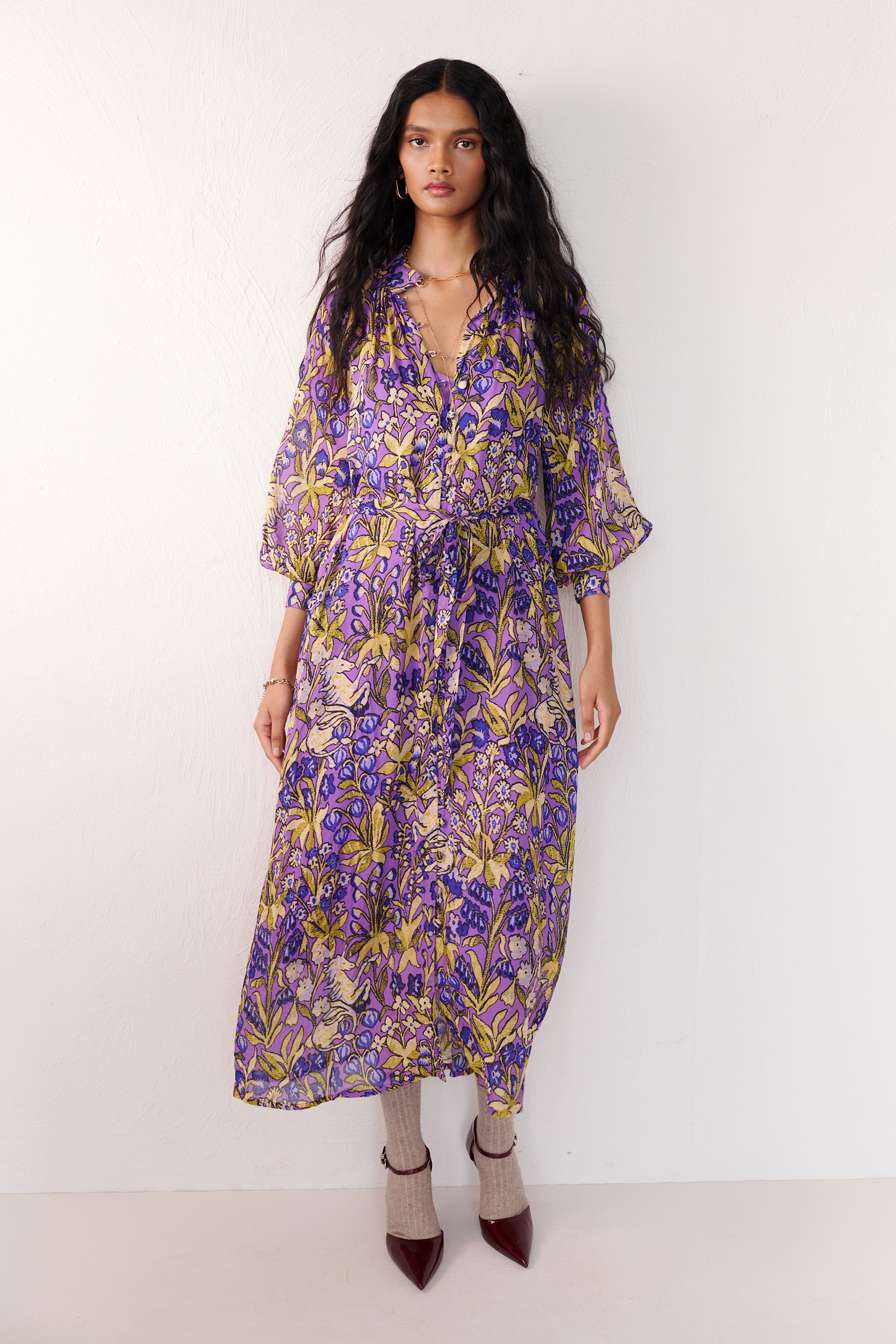 Front view of woman standing. She is wearing the Tara dress in the Unicorn Tapestry print. It is a flowing purple viscose voile dress with a stand collar, gathered neckline and sleeves, fabric-covered buttons, and a removable belt, featuring intricate floral and mythical motifs inspired by the Scottish Highlands.
