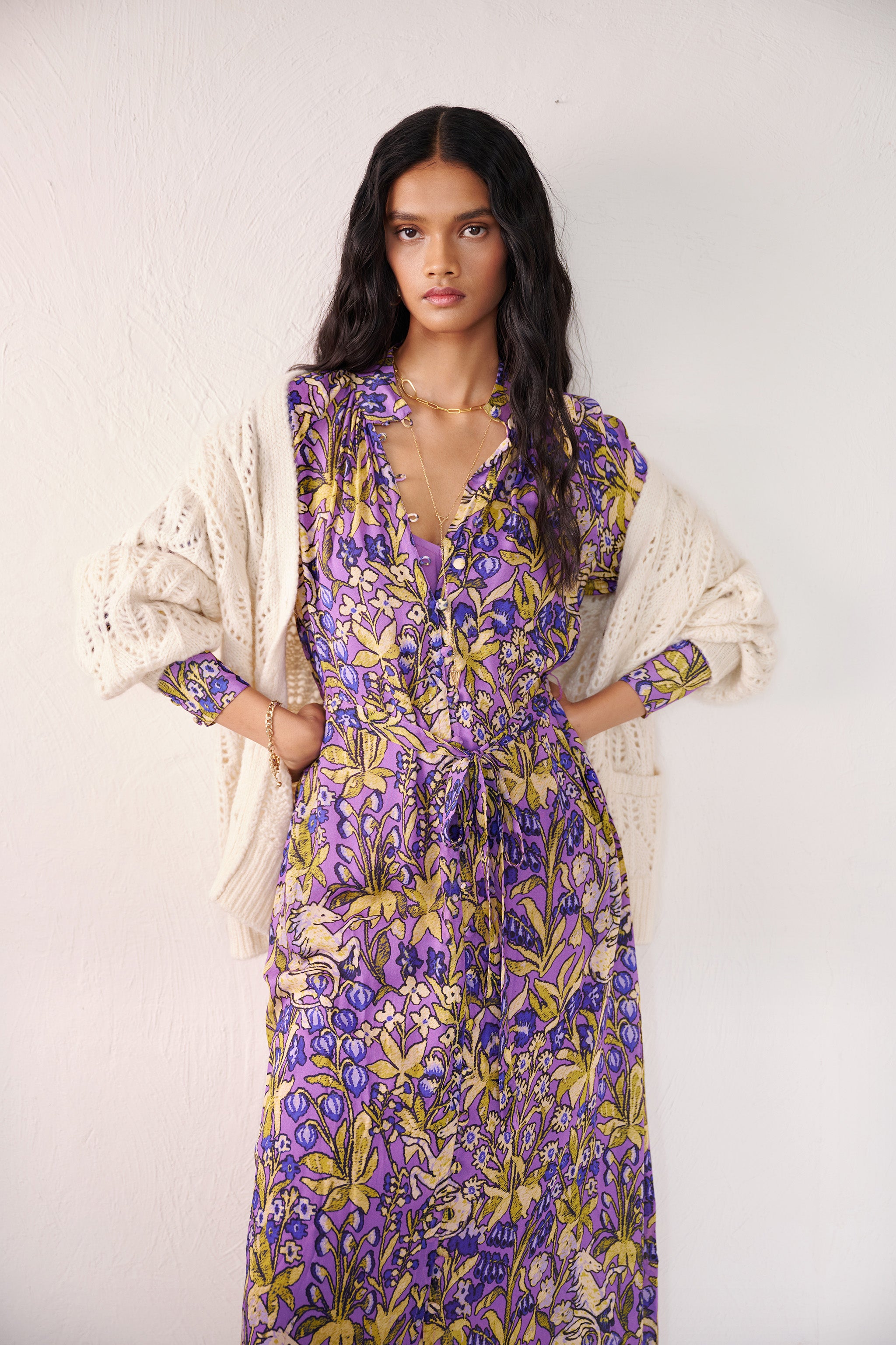 Front view of woman standing. She is wearing the Tara dress in the Unicorn Tapestry print. It is a flowing purple viscose voile dress with a stand collar, gathered neckline and sleeves, fabric-covered buttons, and a removable belt, featuring intricate floral and mythical motifs inspired by the Scottish Highlands. Styled with an ivory boyfriend cardigan.