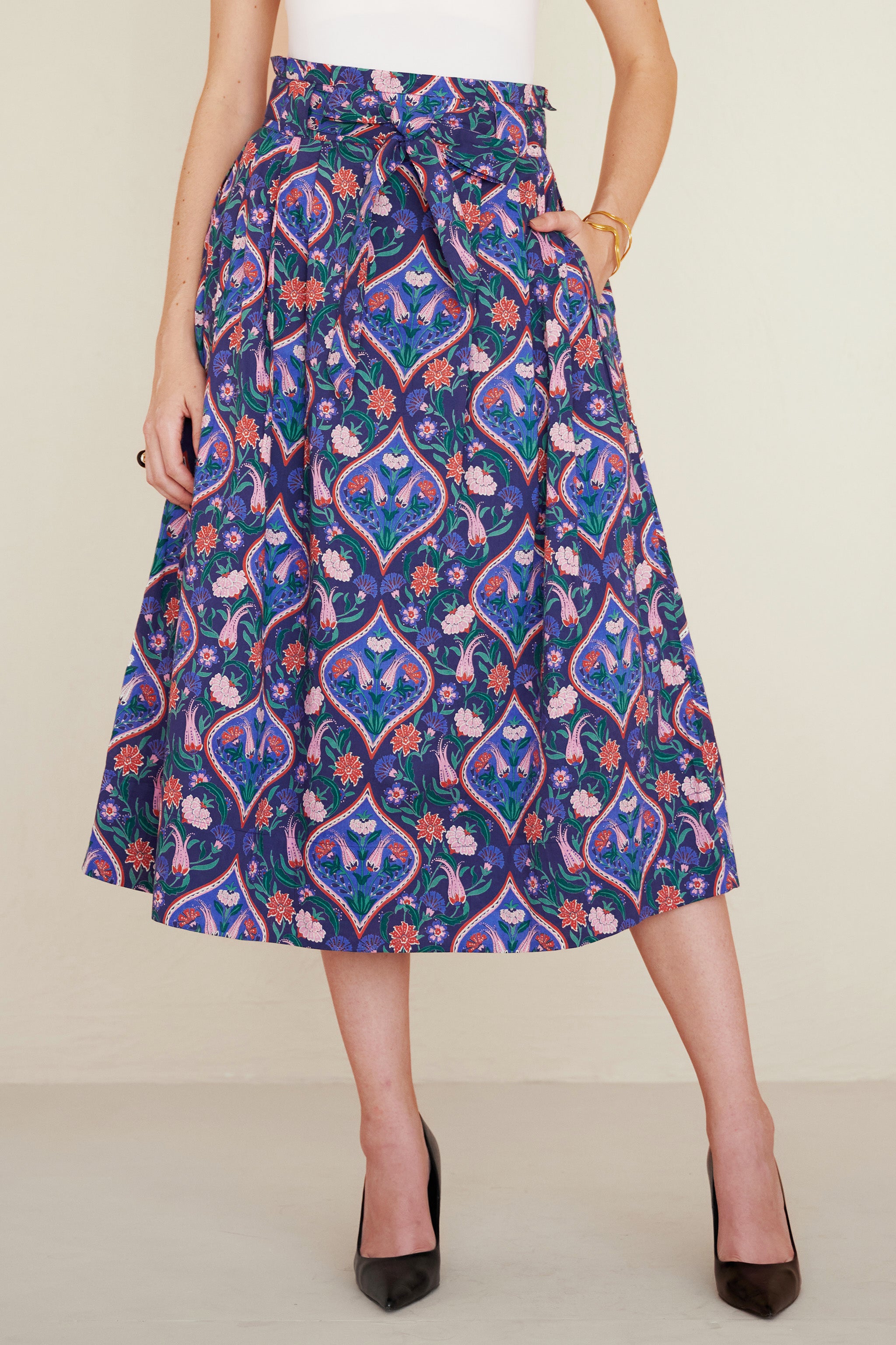 Front view of woman wearing a midi skirt with a paperbag waist, fabric belt, and pockets, featuring a floral tile print inspired by decorative arts.