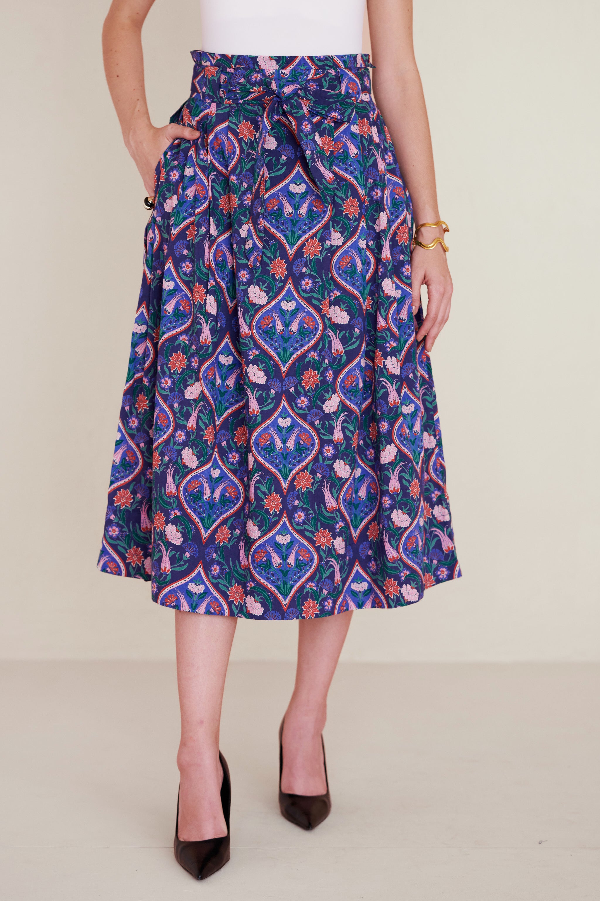 Front view of woman wearing a midi skirt with a paperbag waist, fabric belt, and pockets, featuring a floral tile print inspired by decorative arts.