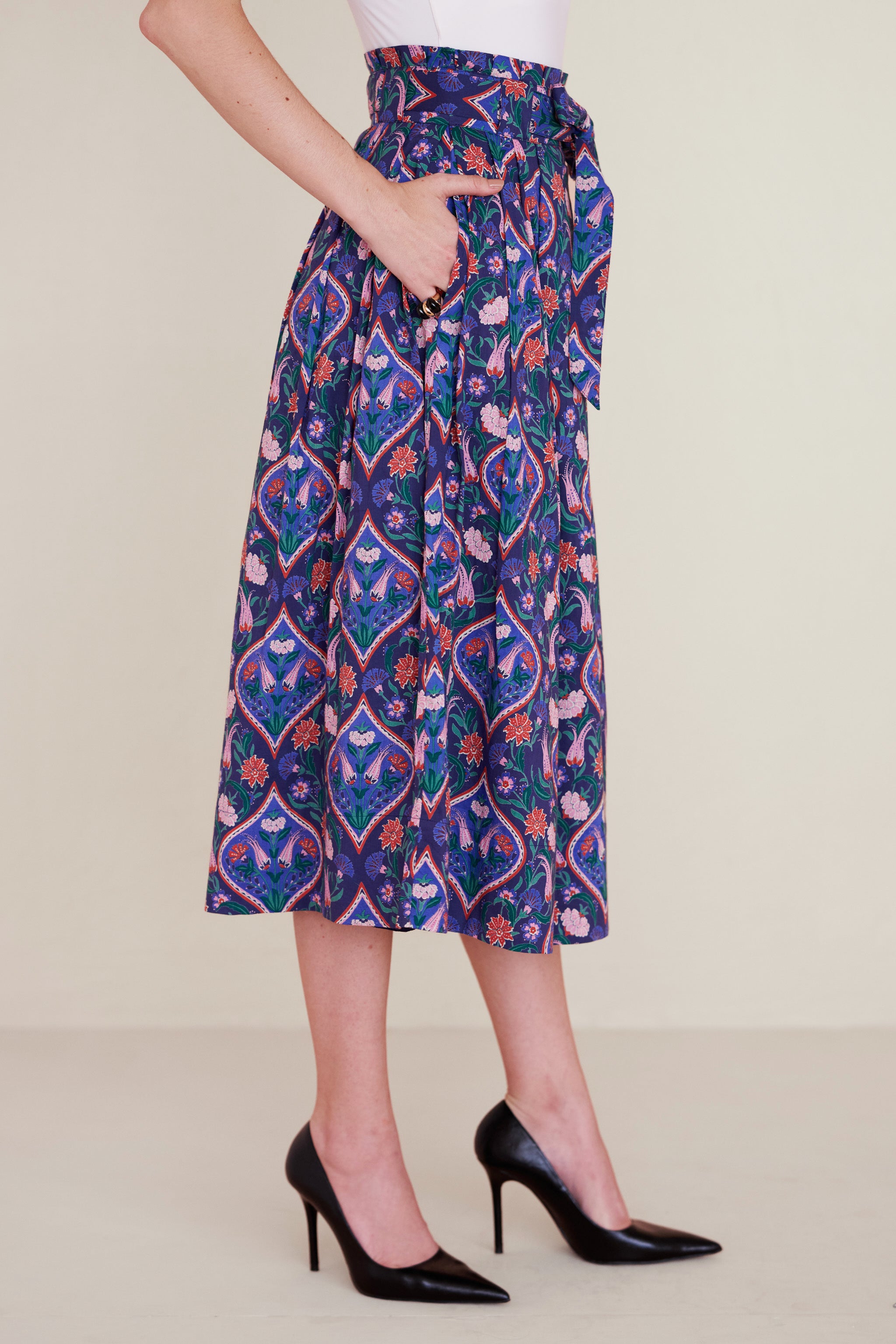 Side view of woman wearing a midi skirt with a paperbag waist, fabric belt, and pockets, featuring a floral tile print inspired by decorative arts.