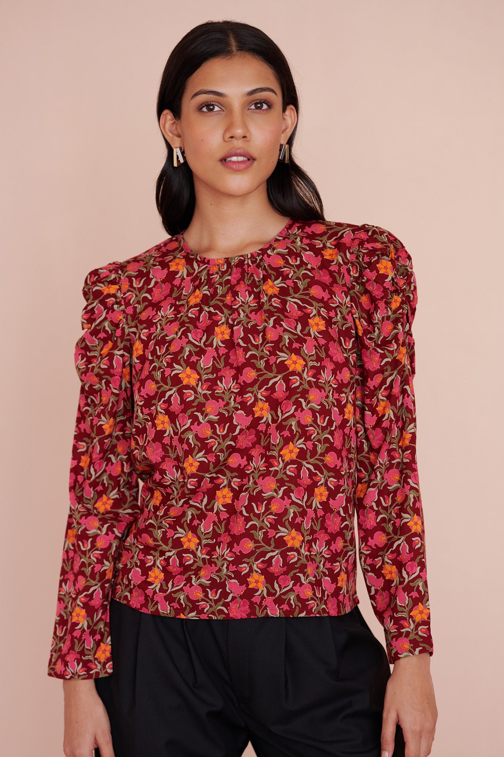 Front view close up of a model standing. She is wearing the Vincent top in eco-friendly viscose, featuring the Pomegranate Vine print in Azalea color. It has soft gathers at the neck and shoulders, long sleeves, and a crew neckline. The elegant day-to-night style fastens with a back zipper.