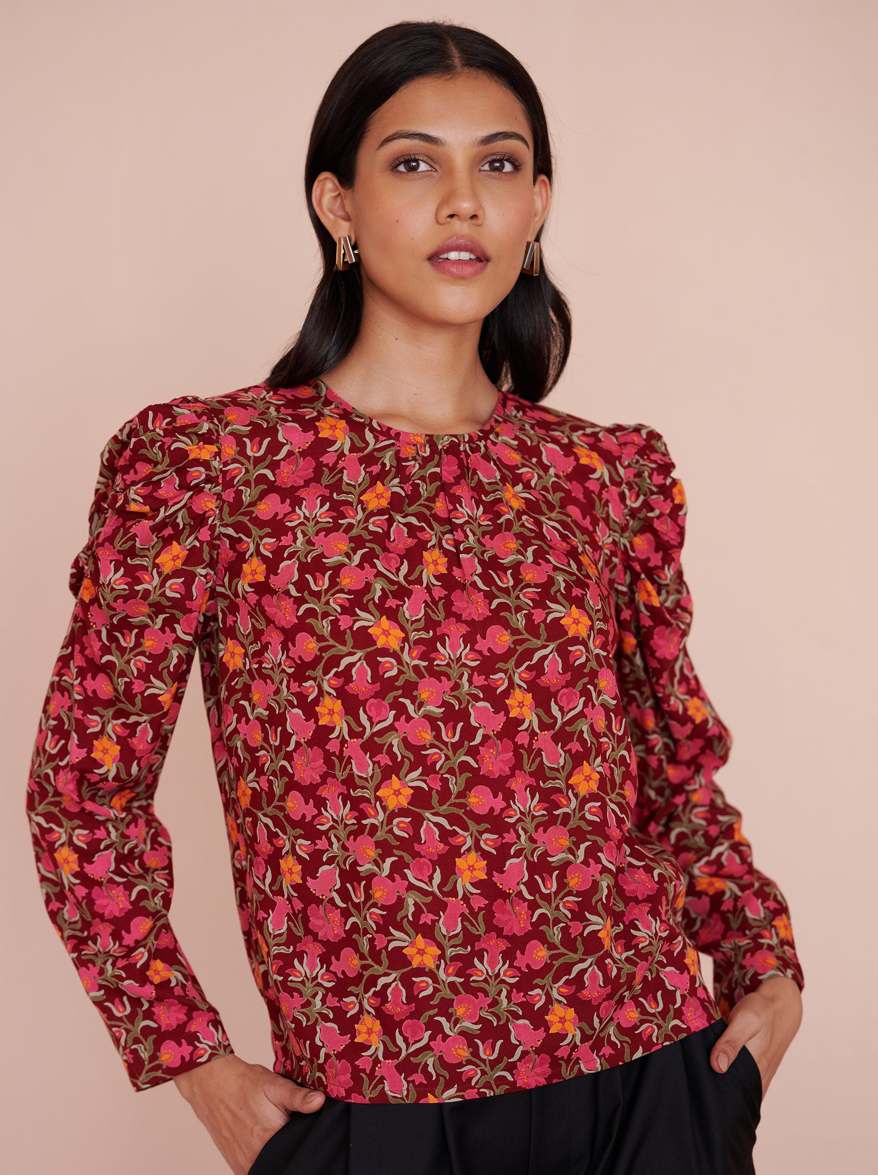 Front view close up of a model standing. She is wearing the Vincent top in eco-friendly viscose, featuring the Pomegranate Vine print in Azalea color. It has soft gathers at the neck and shoulders, long sleeves, and a crew neckline. The elegant day-to-night style fastens with a back zipper.