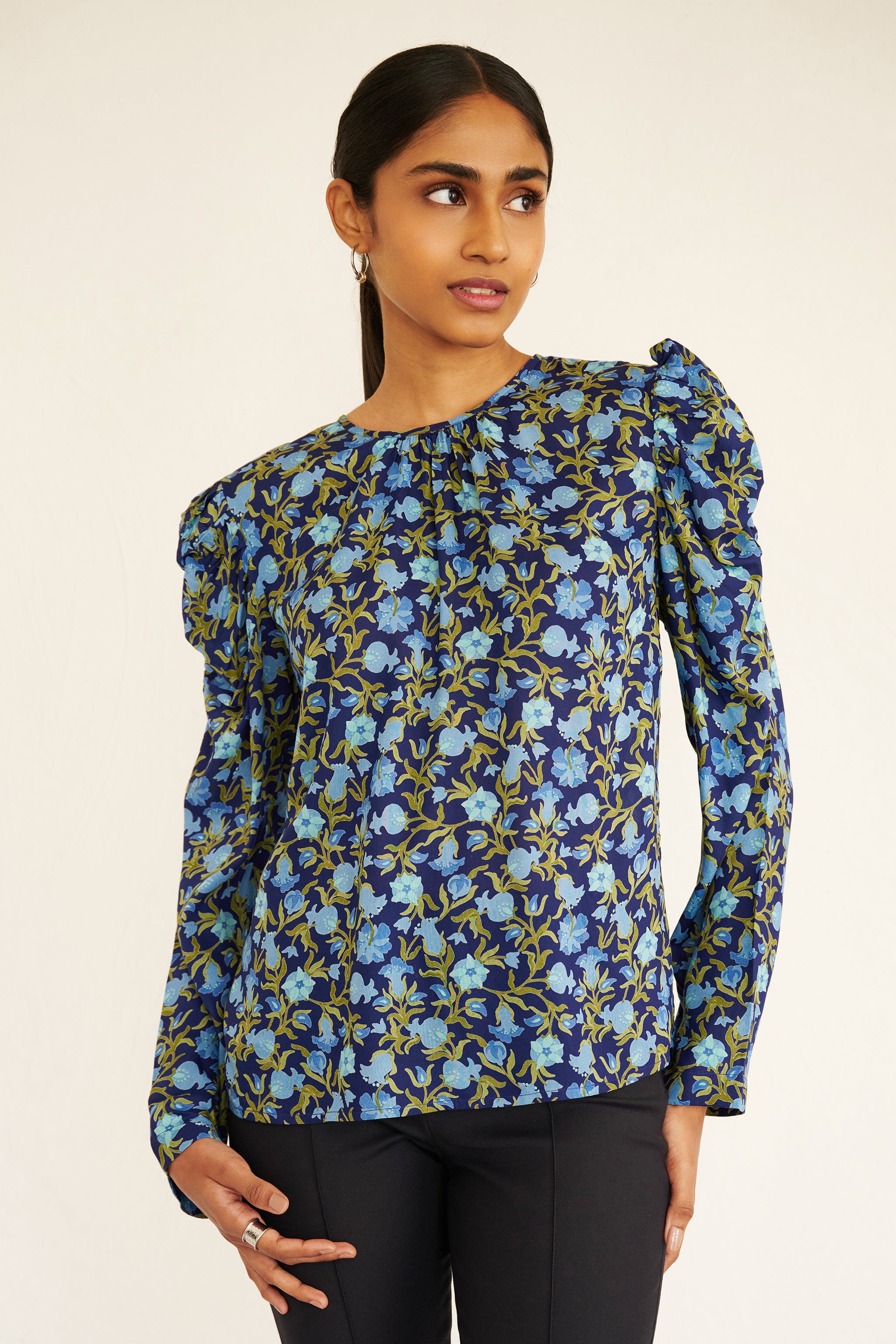 Front view of woman wearing a navy blue blouse with light blue and green pomegranate vine print, featuring soft gathers at the neck and puffed shoulders, paired with slim black trousers and gold hoop earrings.