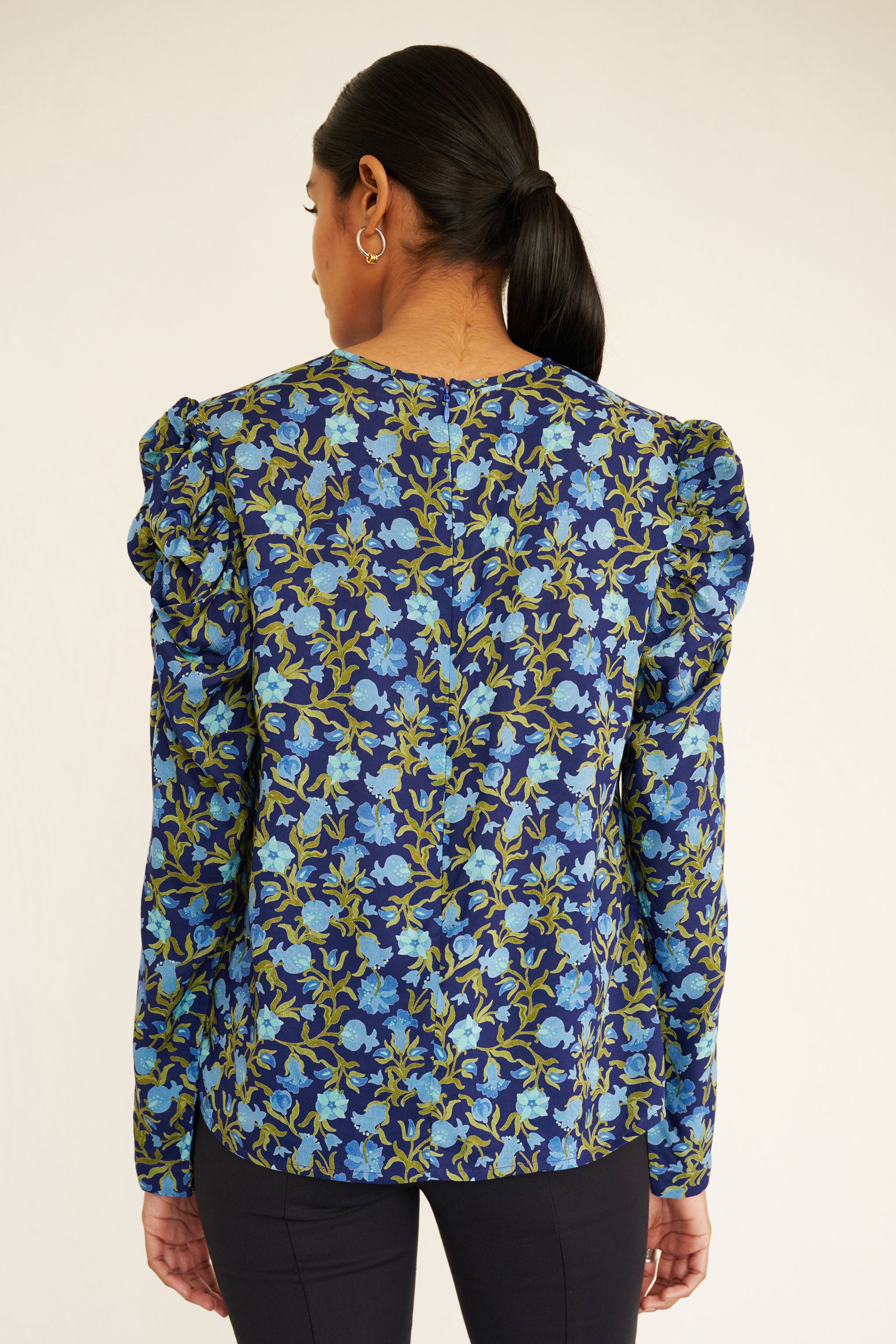 Back view of woman wearing a navy blue blouse with light blue and green pomegranate vine print, featuring soft gathers at the neck and puffed shoulders, paired with slim black trousers and gold hoop earrings.