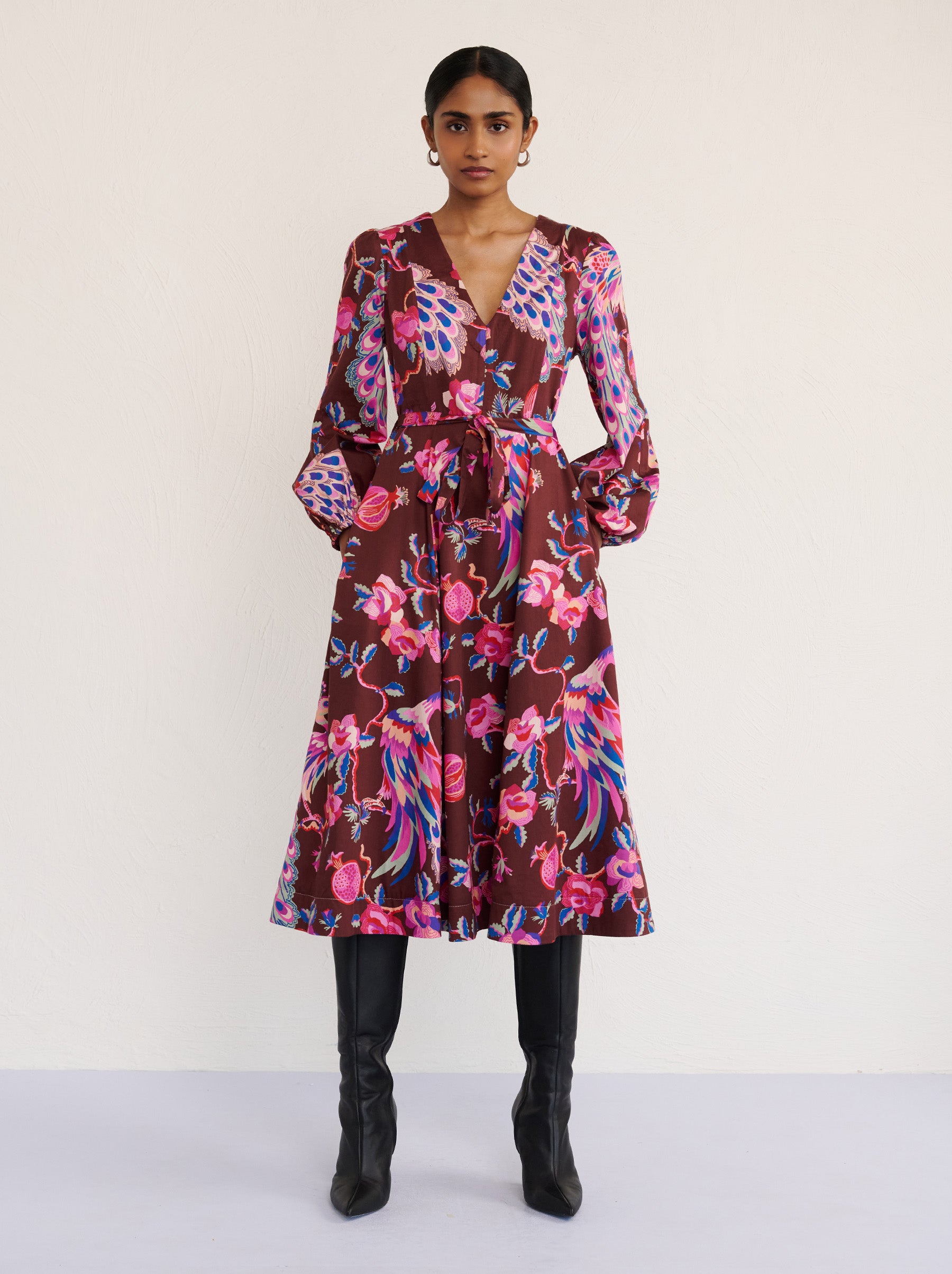 Front view of a model standing. She is wearing the Zora dress in the Bukhara Peacock print in rum color. It is featuring lantern sleeves, a V-neckline, princess seams, a fit-and-flare skirt, a fabric belt, and side-seam pockets.