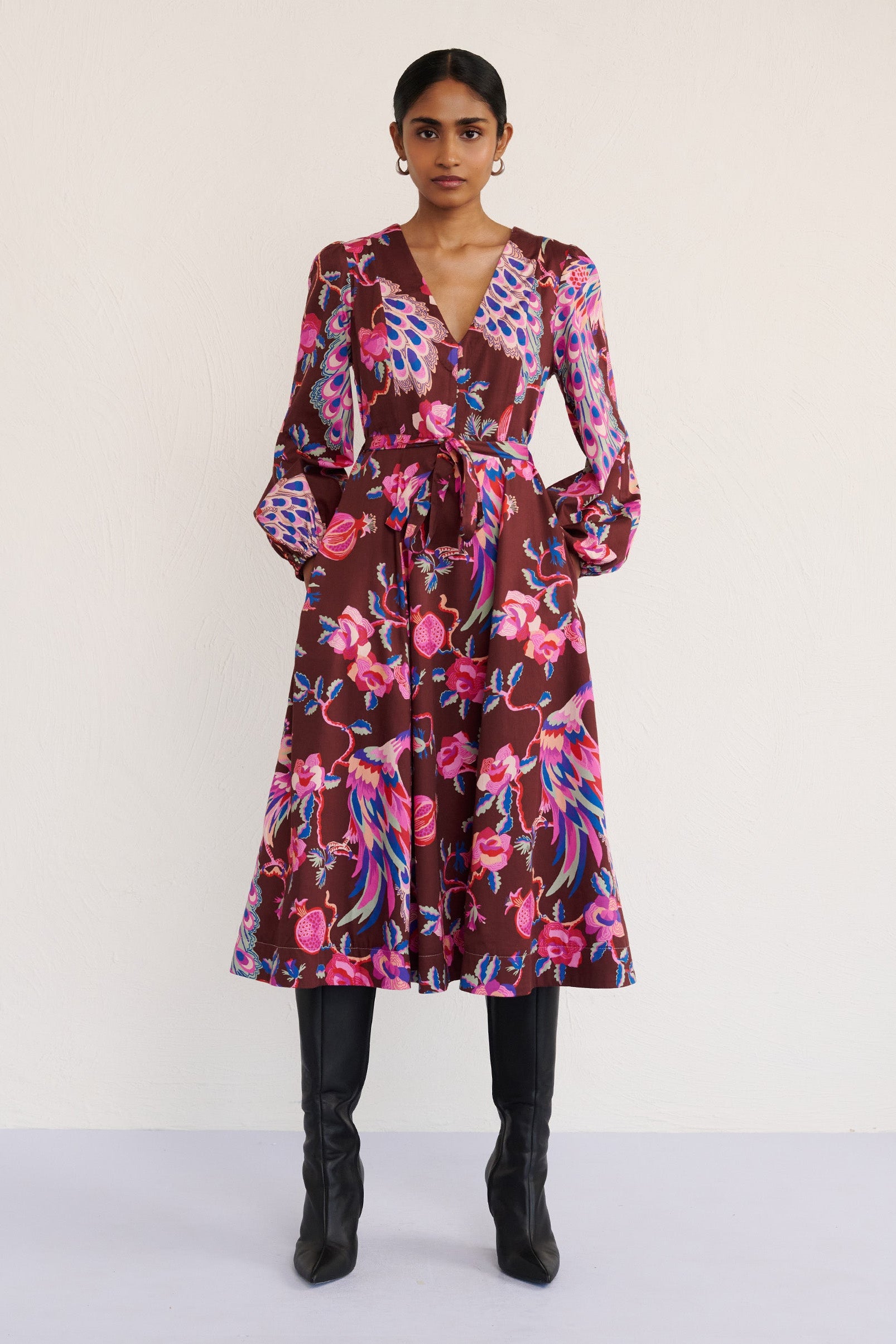 Front view of a model standing. She is wearing the Zora dress in the Bukhara Peacock print in rum color. It is featuring lantern sleeves, a V-neckline, princess seams, a fit-and-flare skirt, a fabric belt, and side-seam pockets.