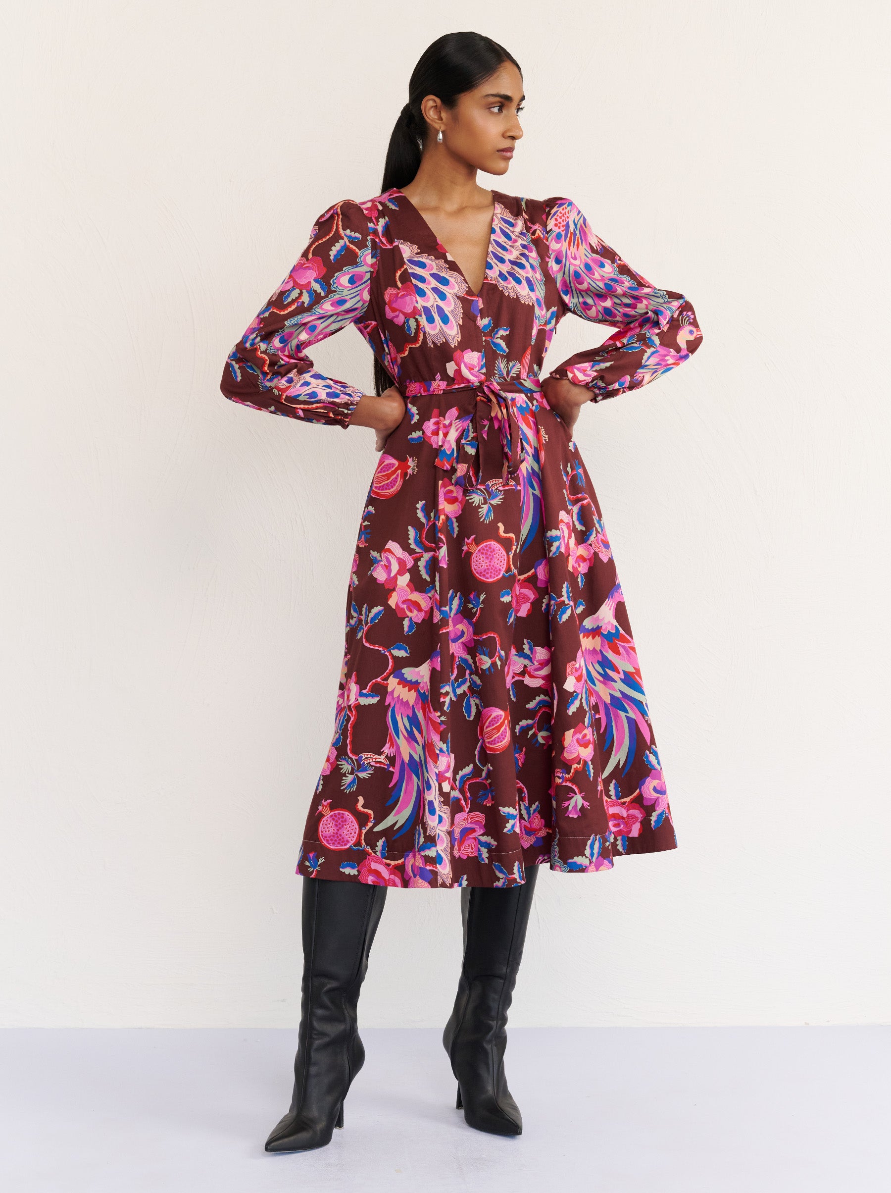 Front view of a model posing. She is wearing the Zora dress in the Bukhara Peacock print in rum color. It is featuring lantern sleeves, a V-neckline, princess seams, a fit-and-flare skirt, a fabric belt, and side-seam pockets.