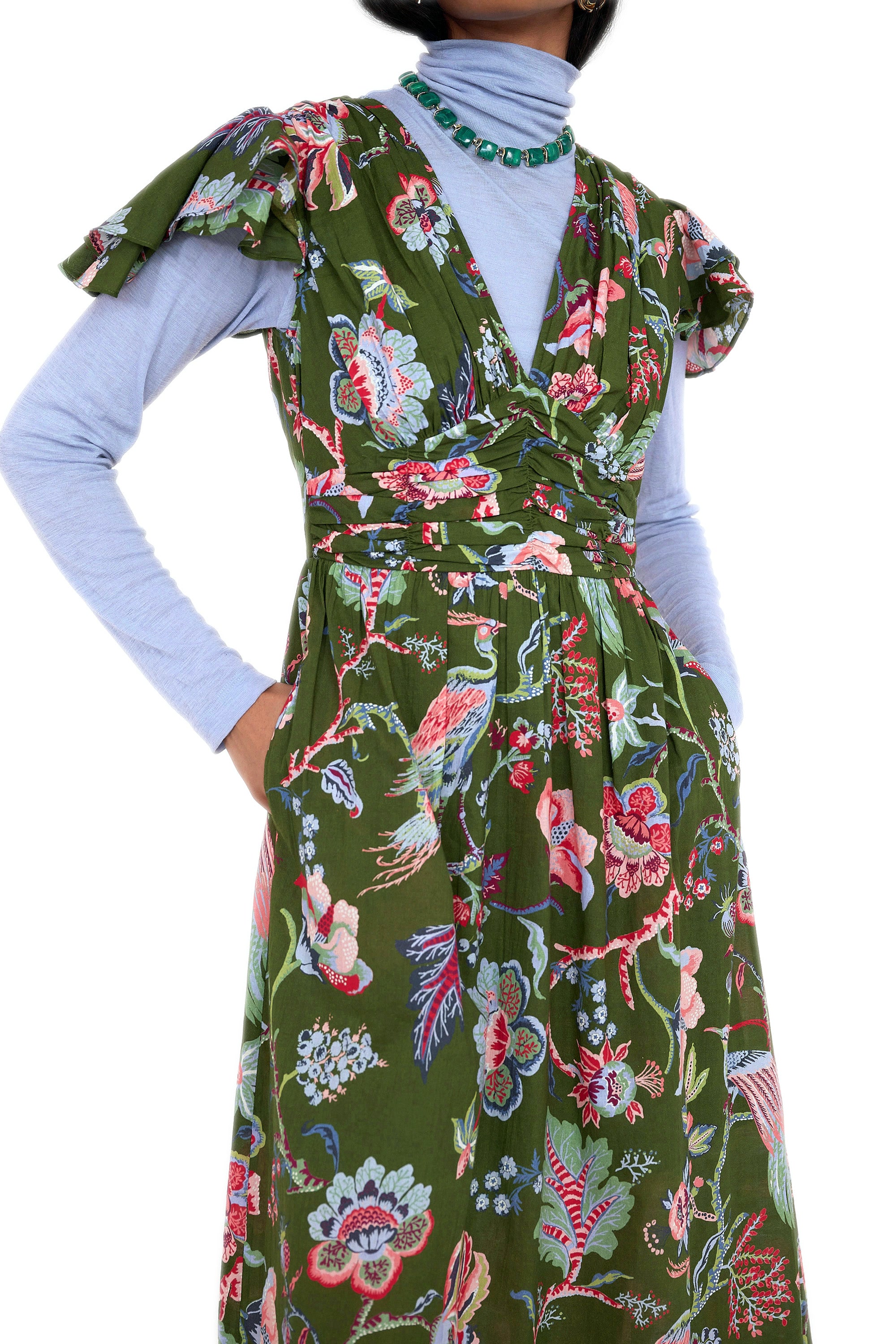 Front view close up of model standing. She is wearing the Adele dress in Chintz print moss color. It features a V-neck, pleat details across the bust, and delicate flutter sleeves. Its styled with a nigh neck layer inside with boots and accessories.