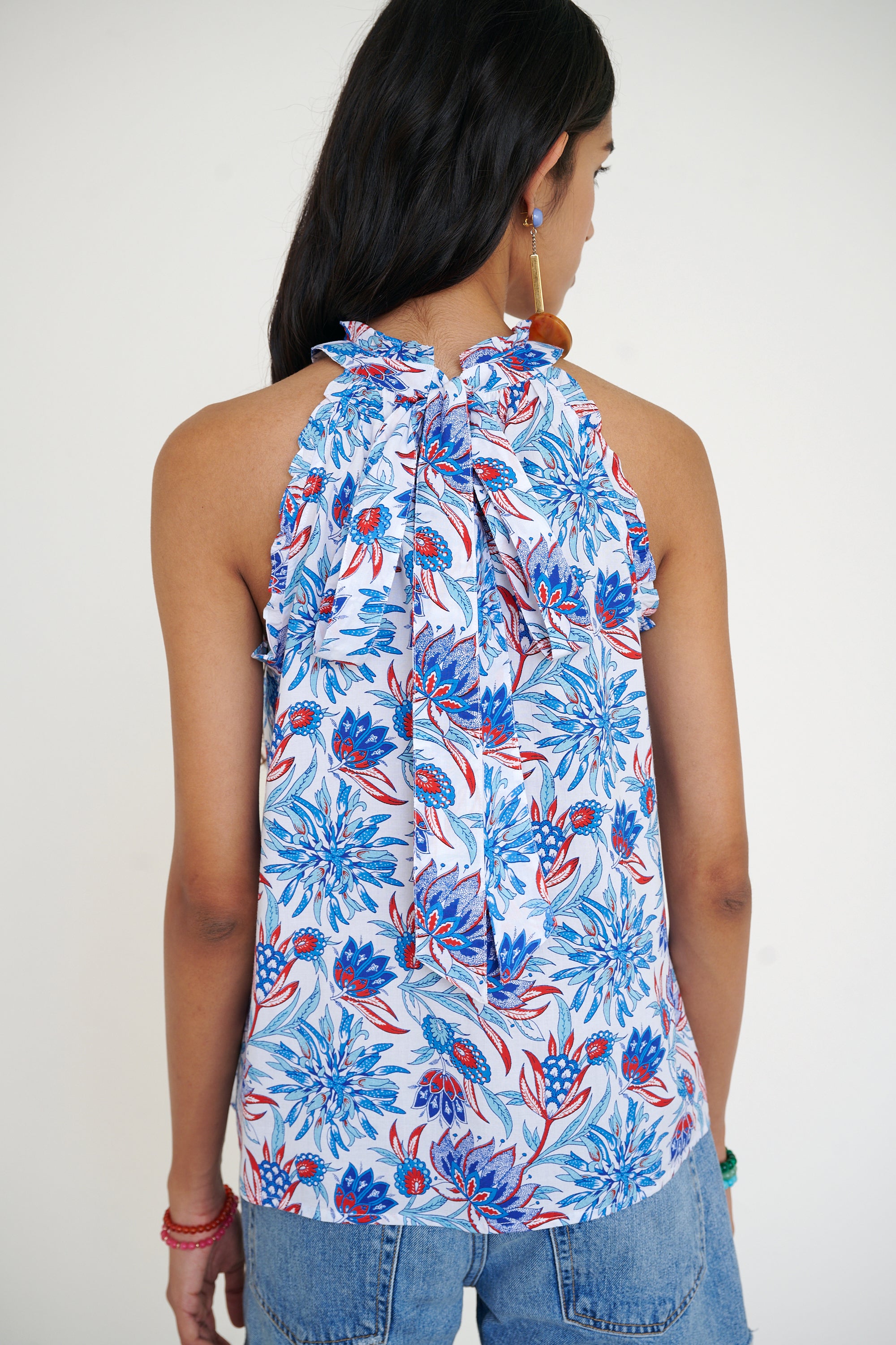 Back view of woman standing wearing the Amy top in the Philippa print, featuring a sleeveless silhouette, subtle ruffle neckline, and a red, white, and blue wildflower pattern. Paired with denim shorts.