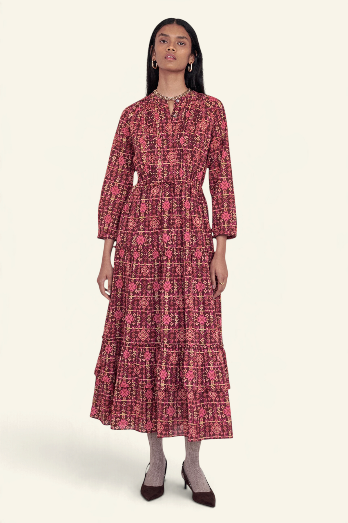 Front view of model standing. She is wearing the Bazaar dress in Needlepoint print, featuring long sleeves, a tiered ankle-length A-line skirt, drawstring belt, and side-seam pockets.