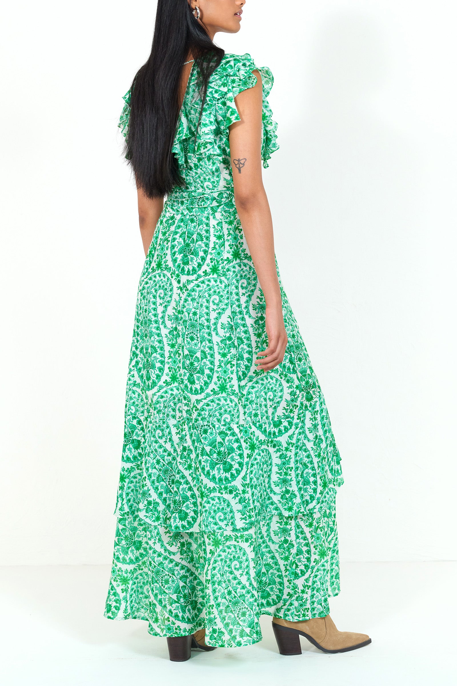 Side view of a woman standing. She is wearing the Beatrice dress in Paisley print, featuring a full-length tiered skirt, ruffled bodice, fabric tie belt, dramatic tie-back neckline, and lustrous cotton silk.