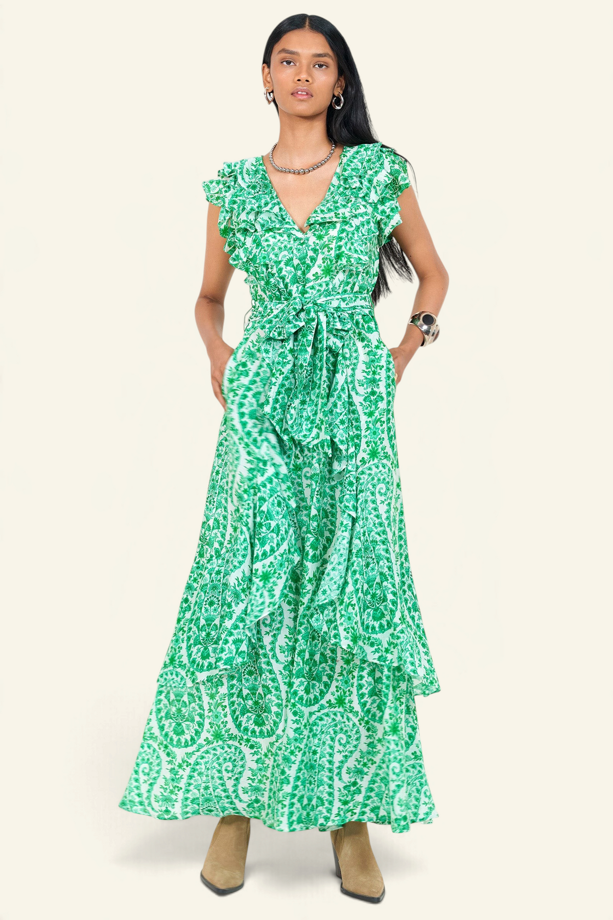 Front view of a woman standing. She is wearing the Beatrice dress in Paisley print with hands in pocket. It features a full-length tiered skirt, ruffled bodice, fabric tie belt, dramatic tie-back neckline, and lustrous cotton silk.