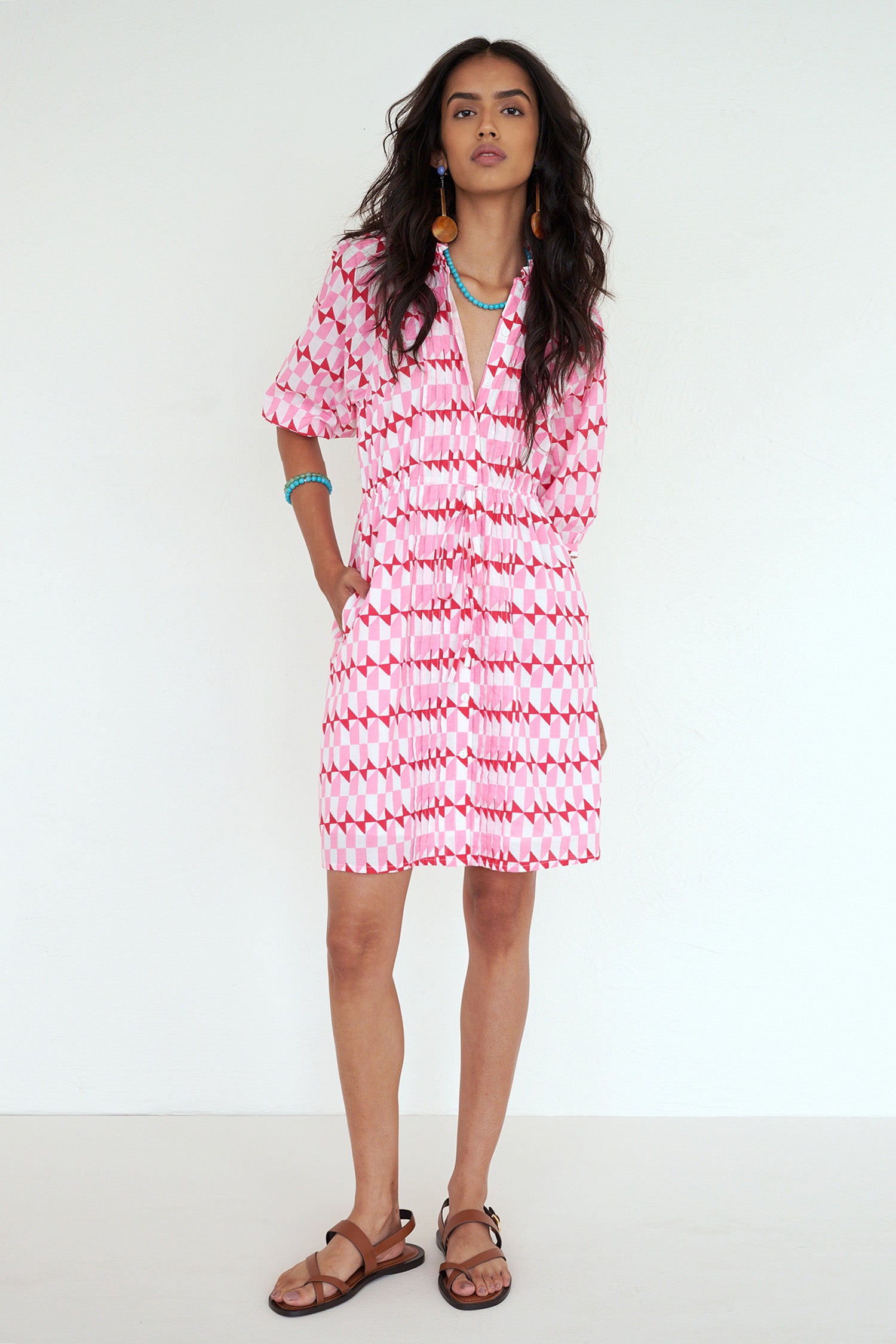 Front view of woman standing. She is wearing the Benita dress in the Stevie print, featuring a shirtdress silhouette, button-front placket, drawstring waist, and geometric motifs inspired by the French Riviera.