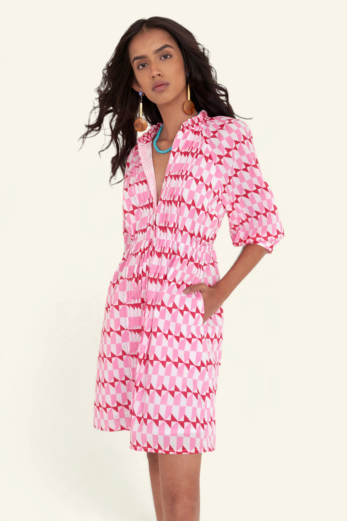 Front side view of woman standing. She is wearing the Benita dress in the Stevie print, featuring a shirtdress silhouette, button-front placket, drawstring waist, and geometric motifs inspired by the French Riviera.