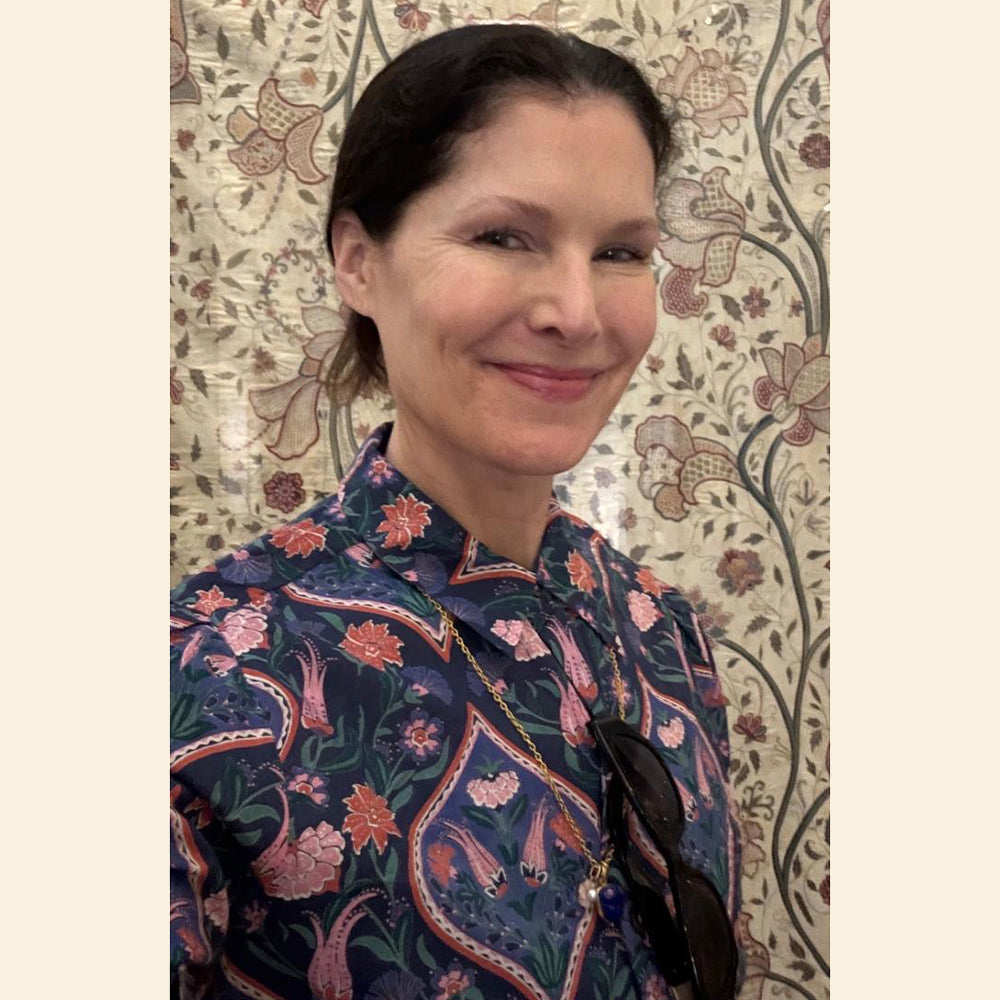 Woman wearing a patterned blouse standing against a decorative wall.
